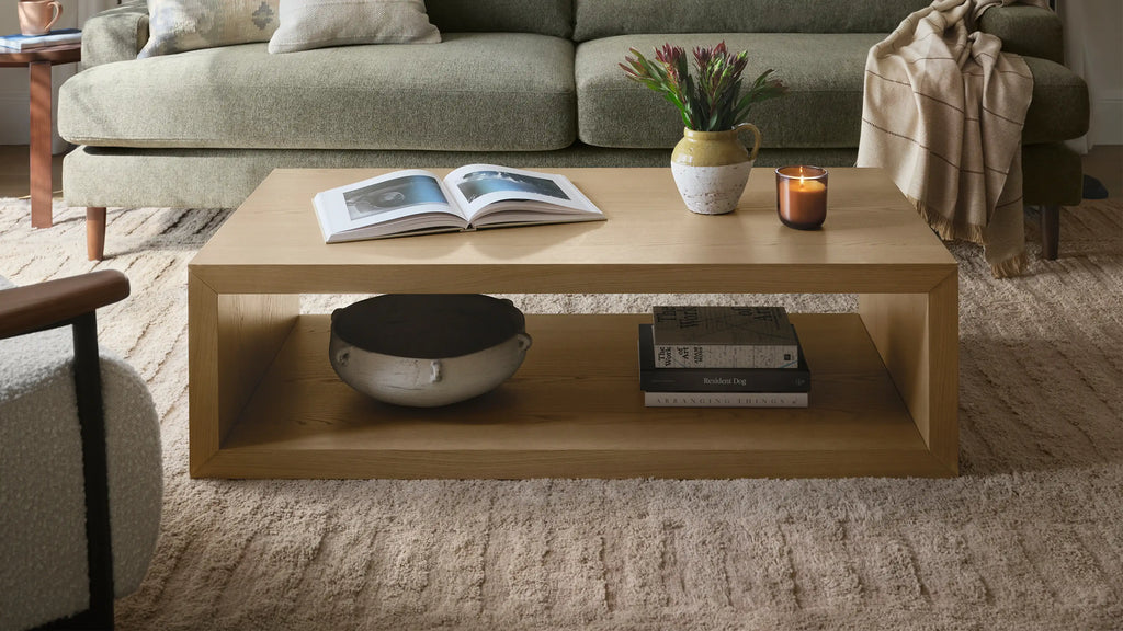 Rectangular wooden coffee table with open shelf, decorated with an open book, vase with flowers, lit candle, pottery bowl, and stacked books.