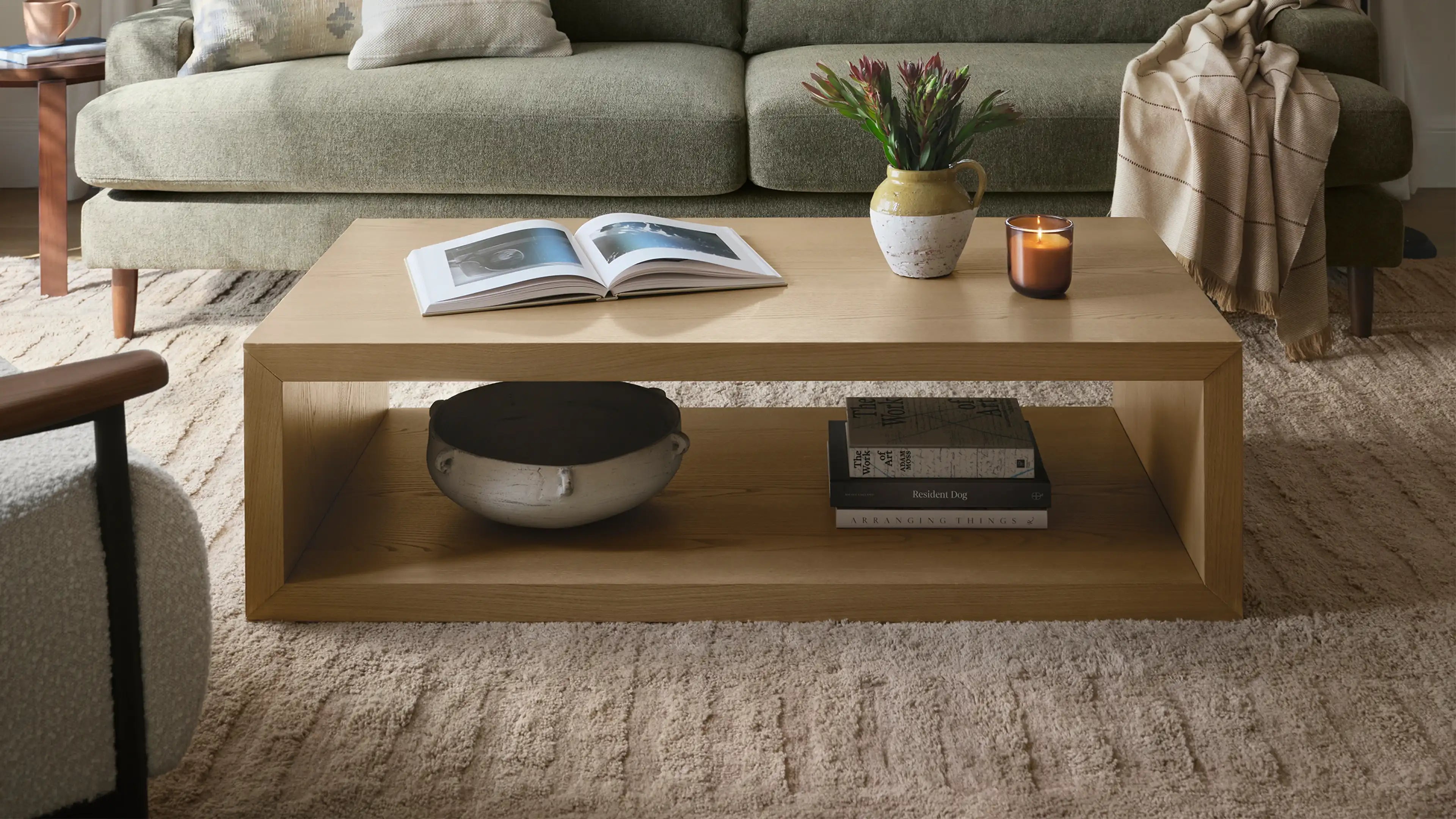 Rectangular wooden coffee table with open shelf, decorated with an open book, vase with flowers, lit candle, pottery bowl, and stacked books.