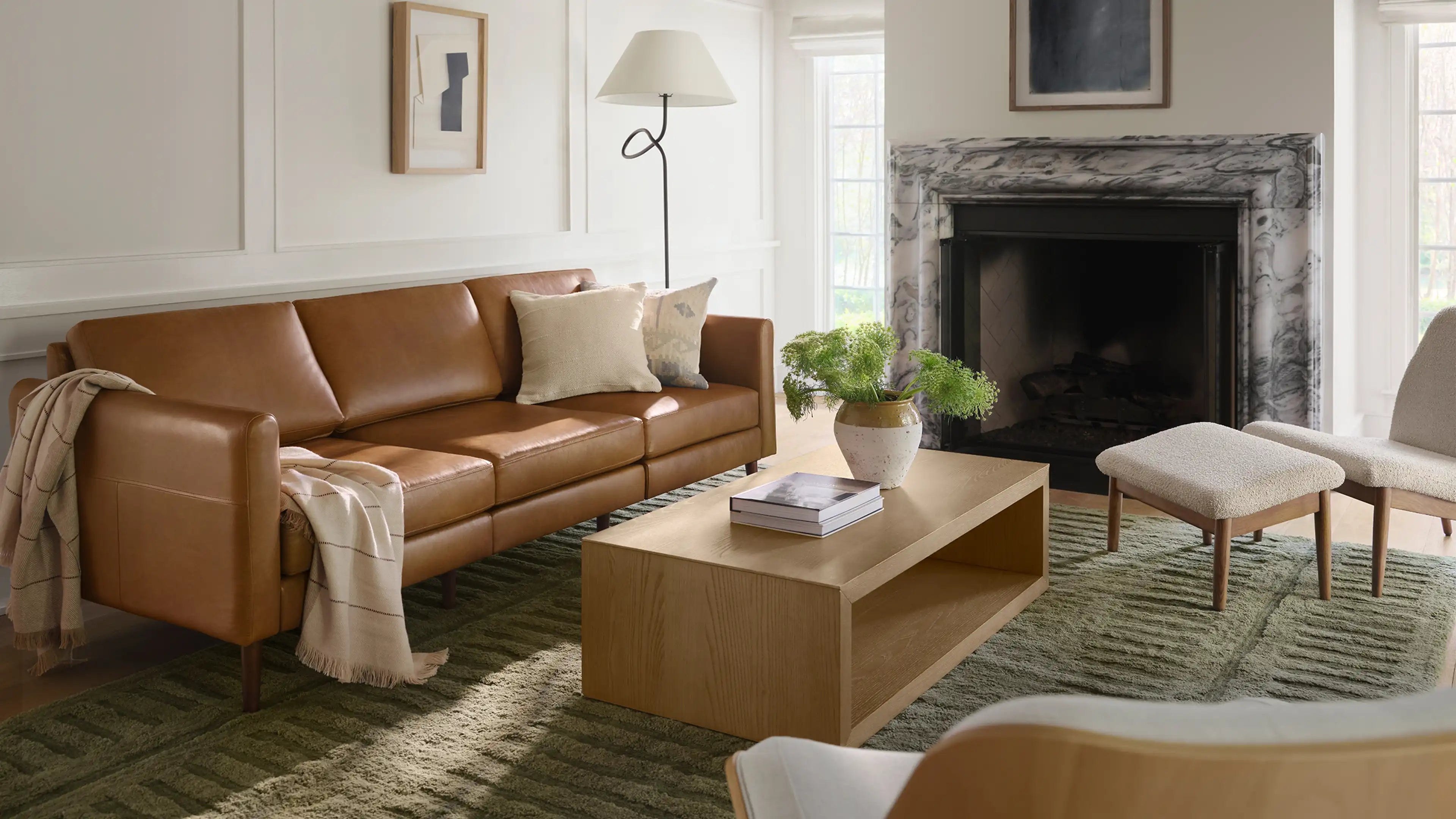 Modern living room with a tan leather sofa, wooden coffee table, white armchair with ottoman, green textured rug, marble fireplace, and floor lamp.
