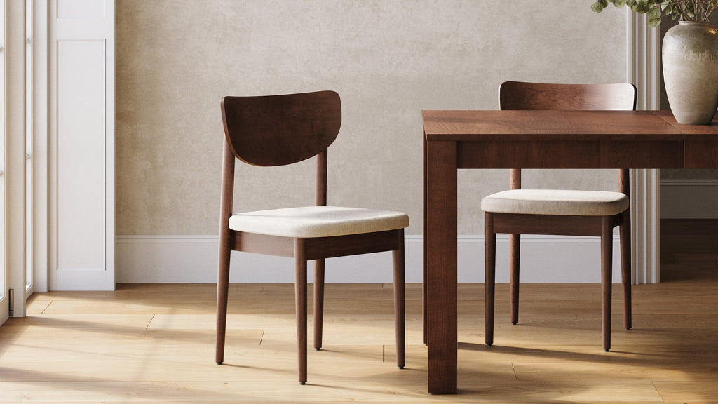 Two modern wooden dining chairs with beige cushioned seats next to a matching wooden dining table in a bright room.