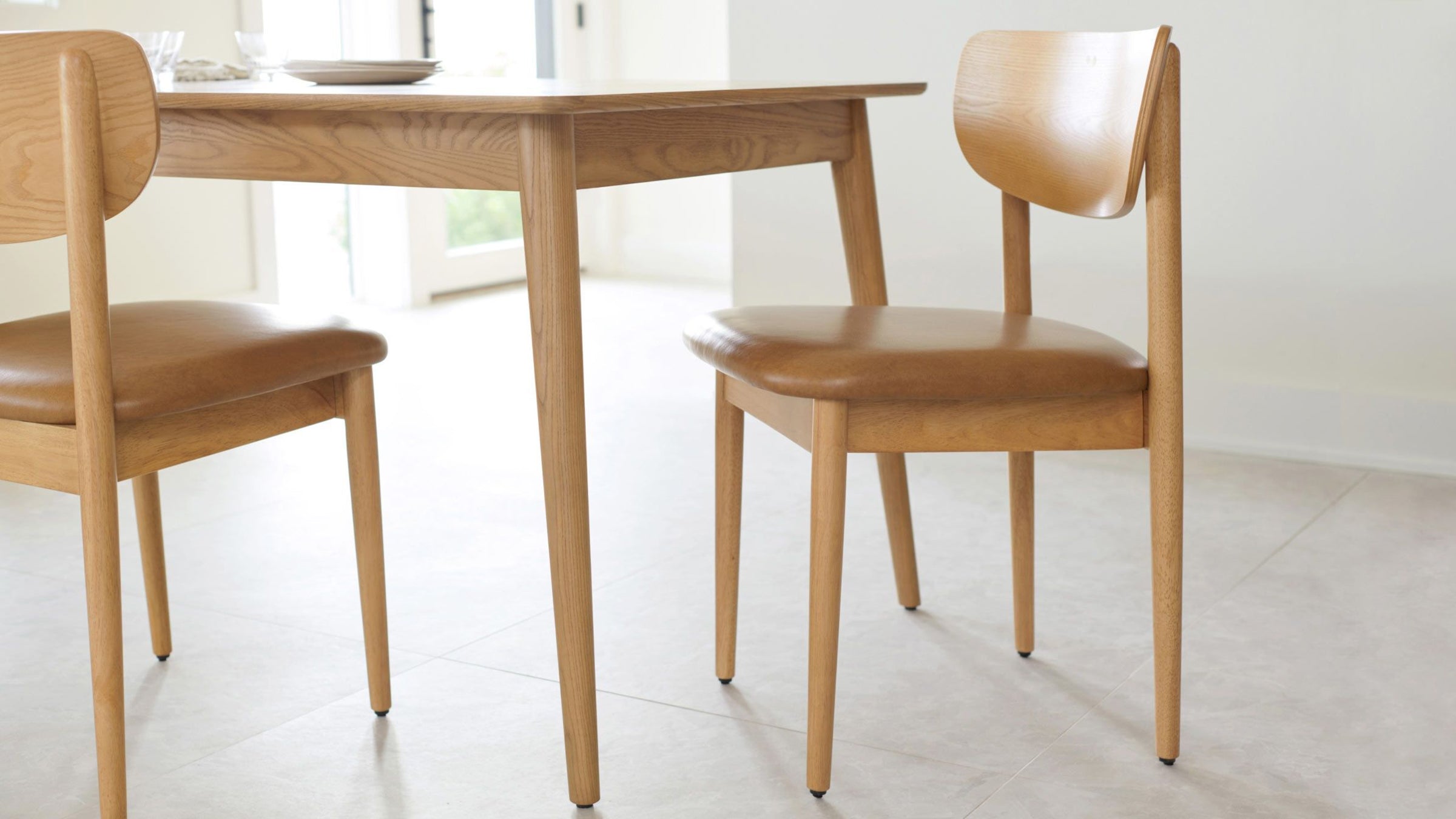 Light wood dining table and matching chairs with tan cushioned seats, set on a light-colored tiled floor in a bright room.