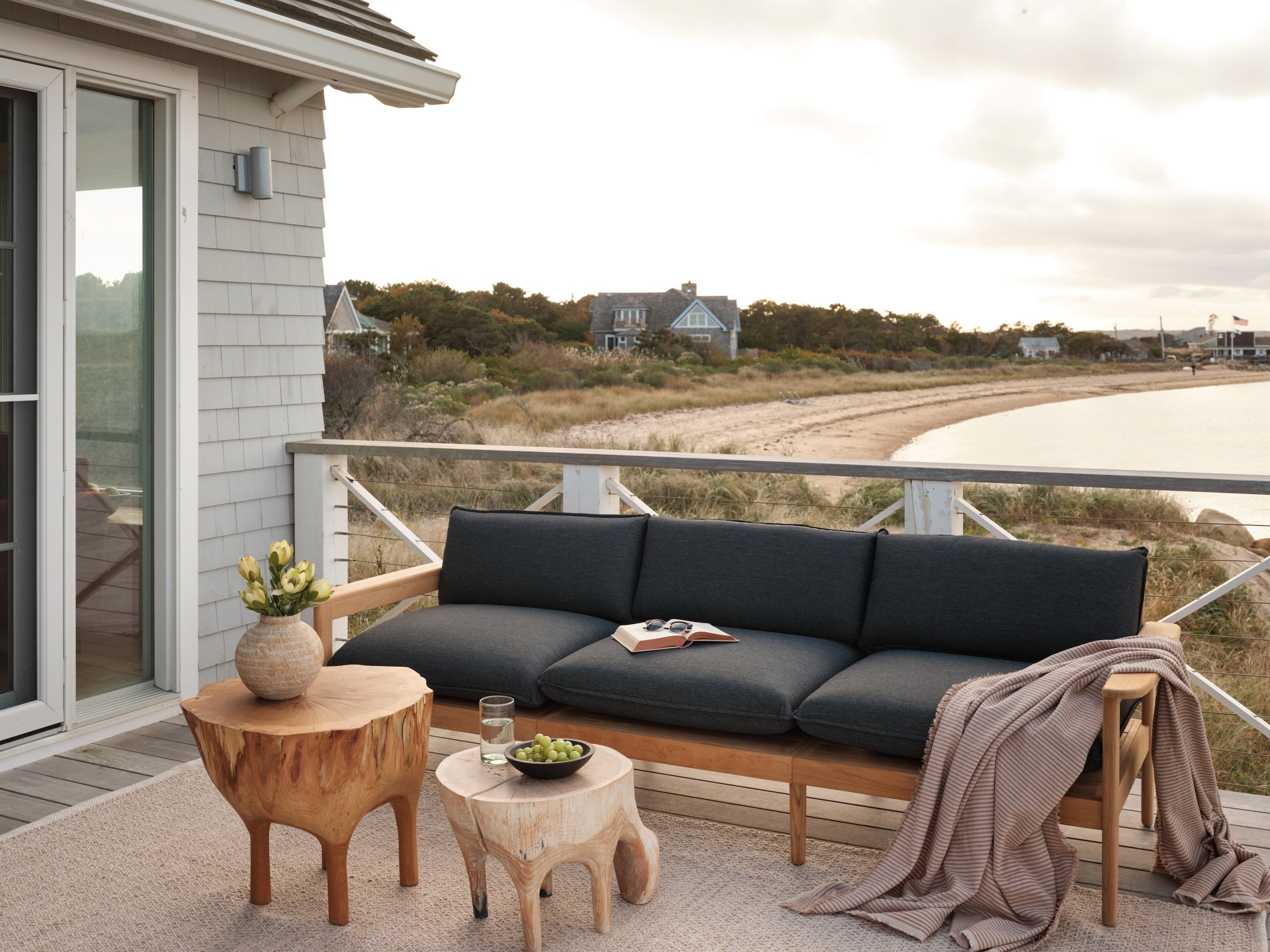 Outdoor seating area on a wooden deck with a charcoal cushion sofa, rustic wooden side tables, vase with flowers, and a coastal shoreline view.