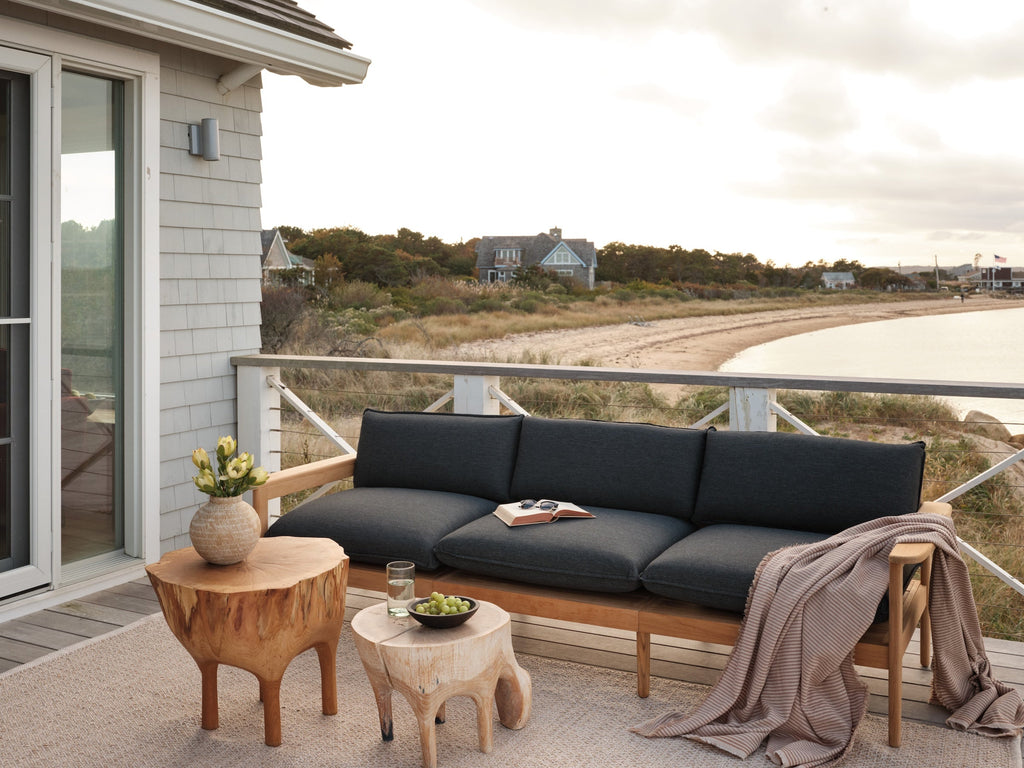 Outdoor seating area on a wooden deck with a charcoal cushion sofa, rustic wooden side tables, vase with flowers, and a coastal shoreline view.