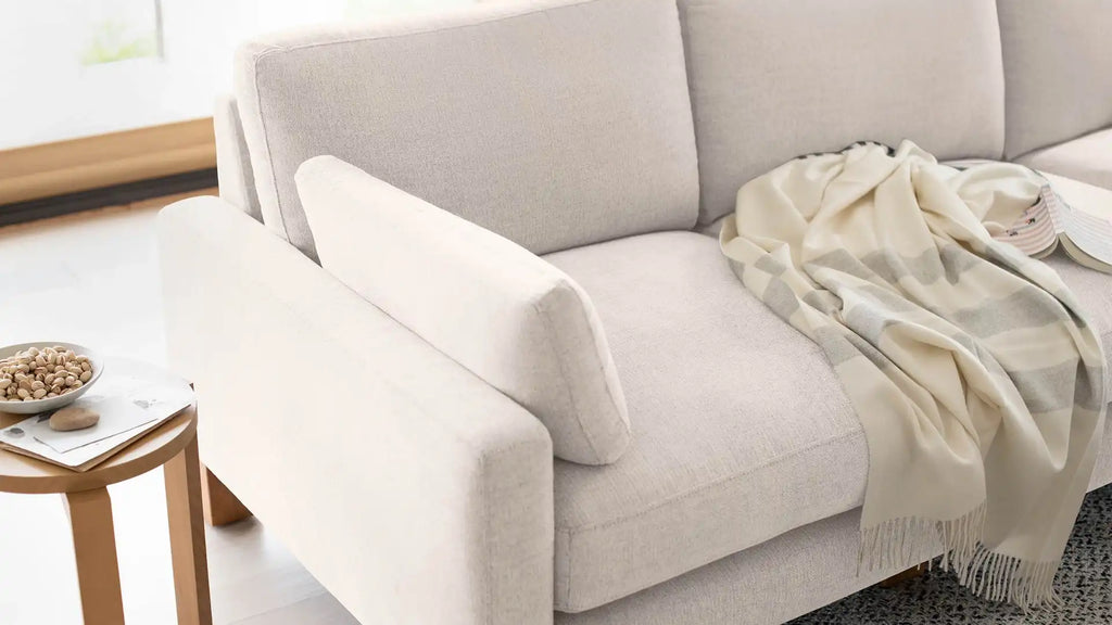 modern beige sofa with a blanket on it and a wooden side table with a bowl of nuts on the side