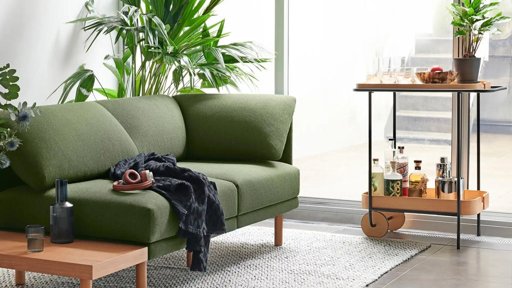 Modern green upholstered sofa with wooden legs next to a wooden coffee table, bar cart with bottles, glasses, and plants.