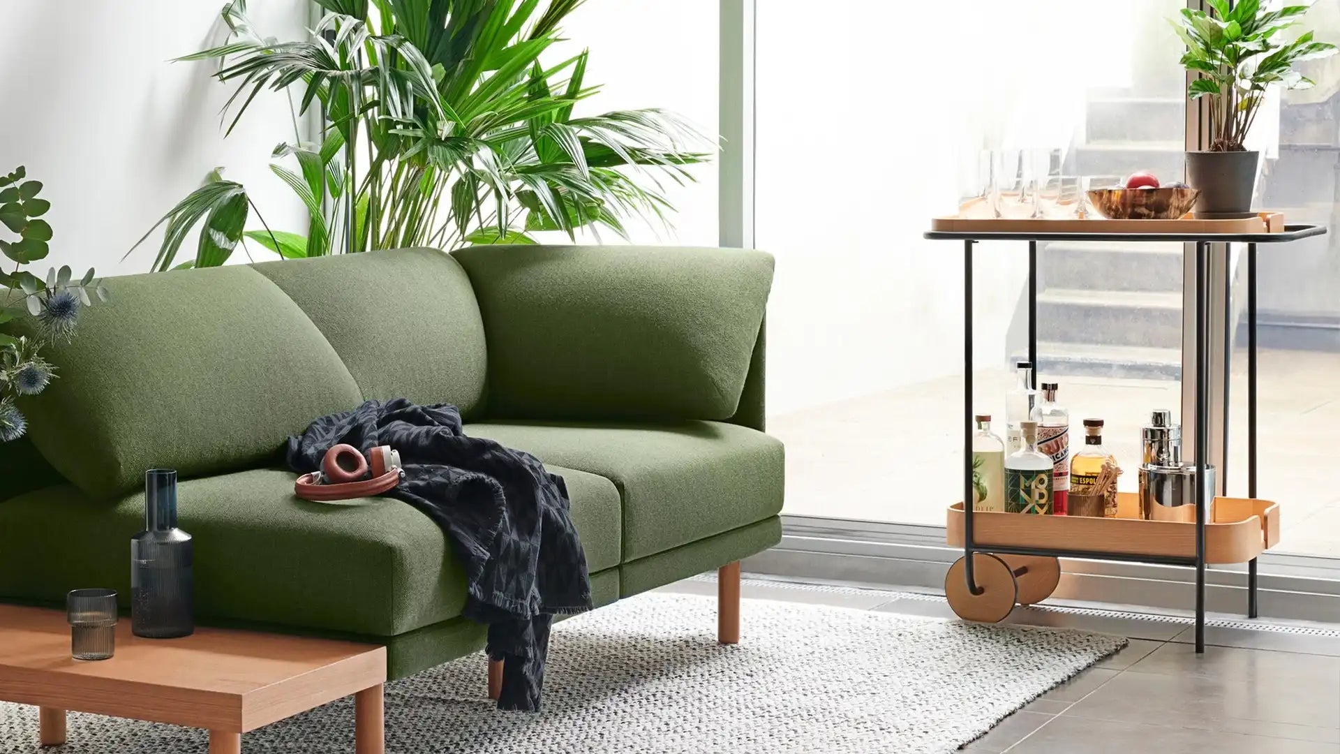 Modern green upholstered sofa with wooden legs next to a wooden coffee table, bar cart with bottles, glasses, and plants.