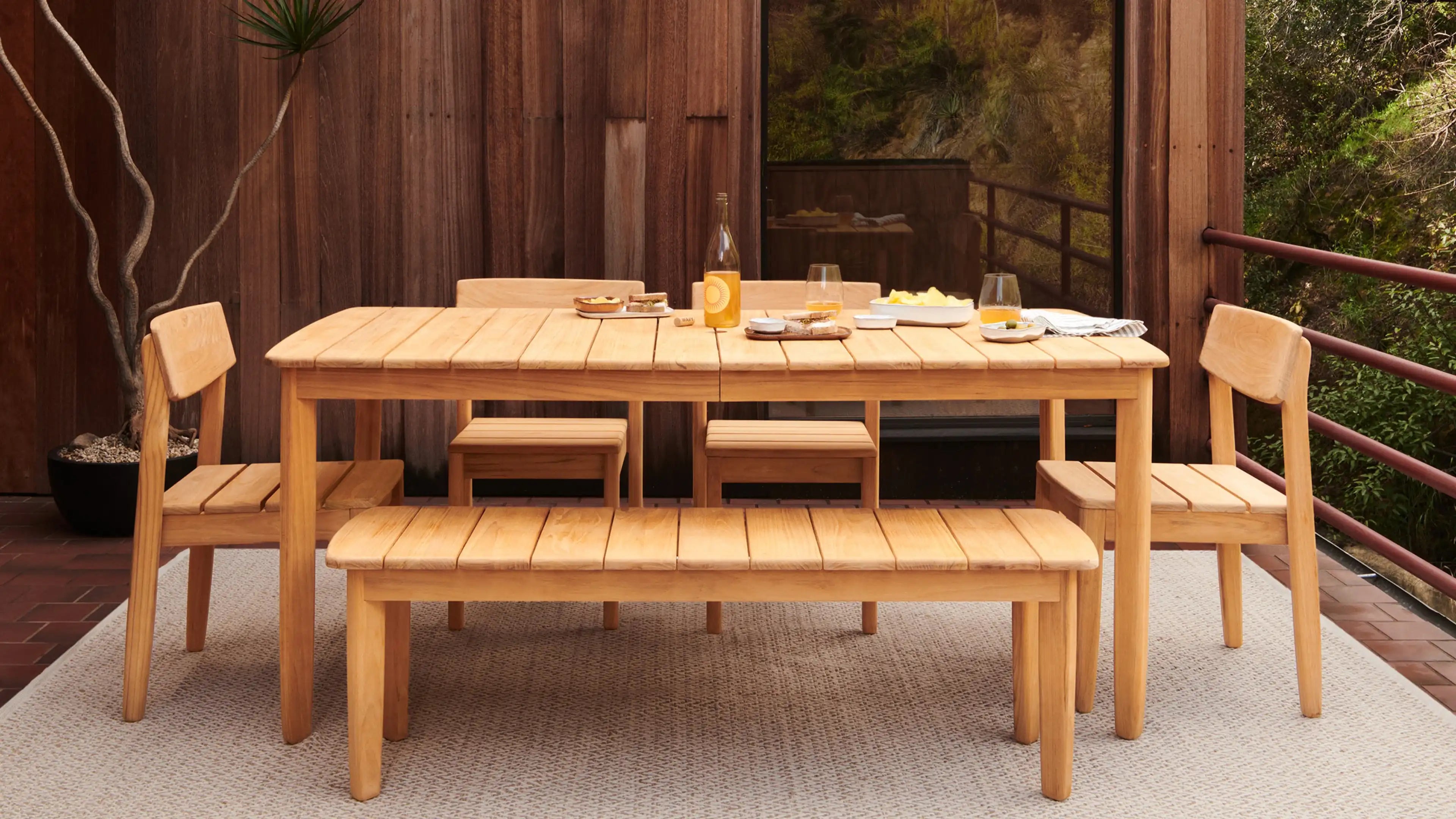 Light wood outdoor dining set with rectangular table, two chairs, two stools, and a bench on a woven rug.