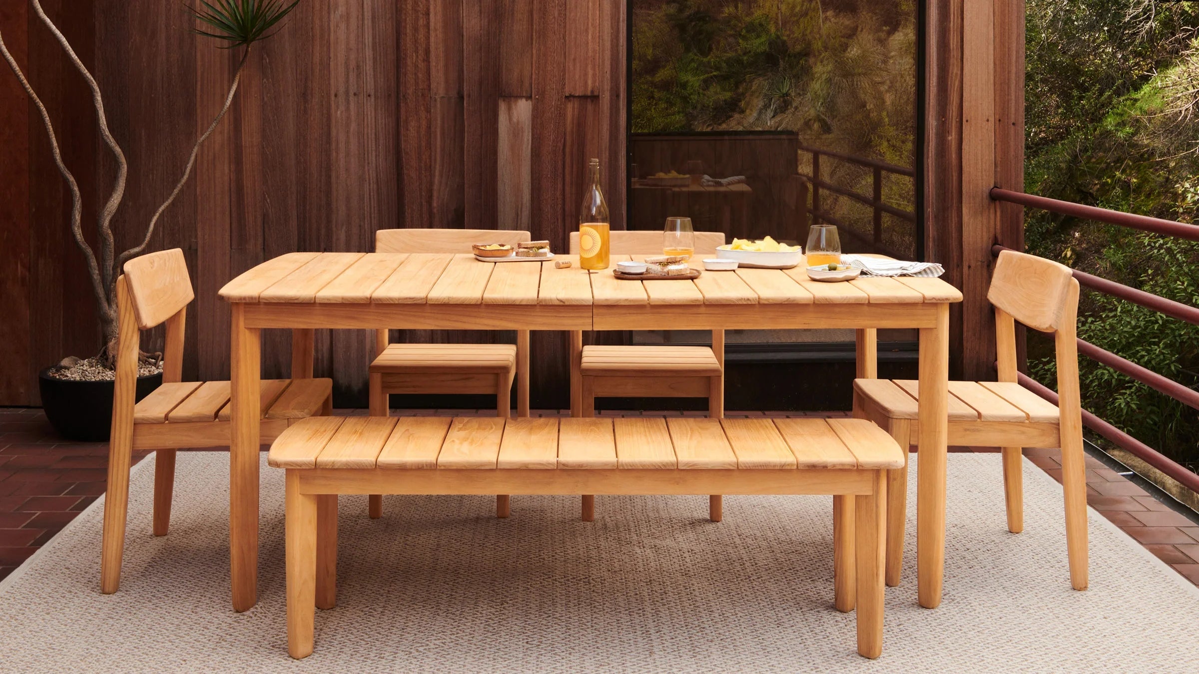 Wooden outdoor dining set with table, bench, and four chairs on a beige rug, decorated with food and drinks.