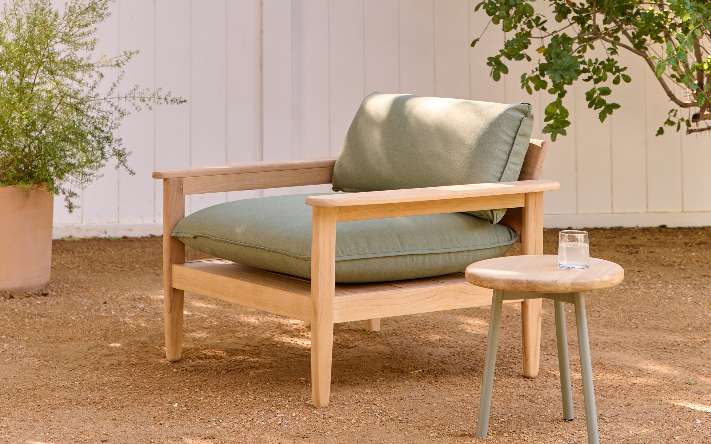 Wooden outdoor armchair with green cushions, placed on dirt ground beside a small round wooden table with a glass.