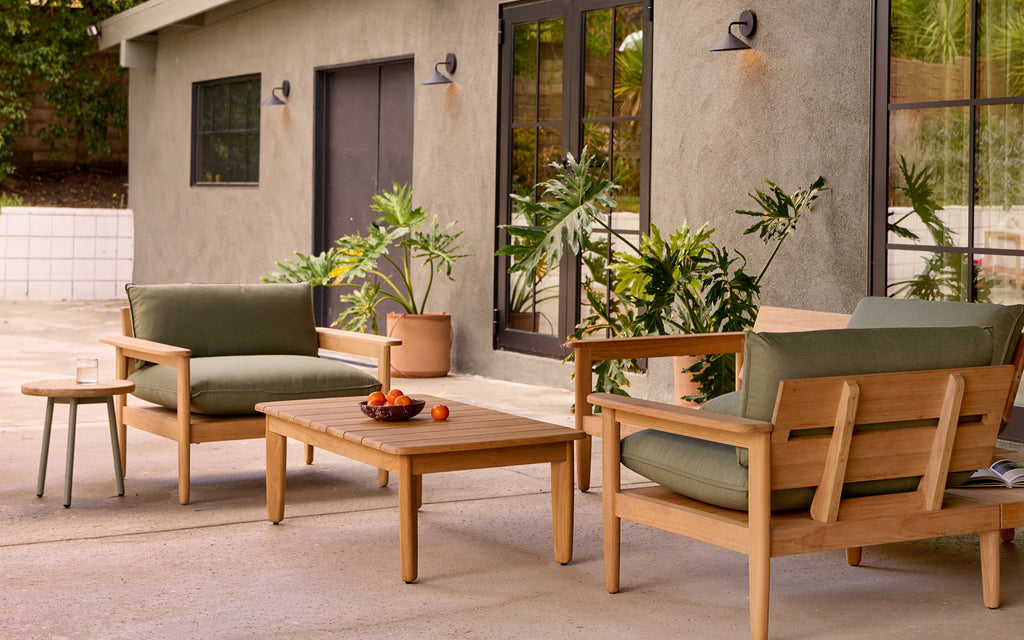 Outdoor patio furniture set with wooden frame, green cushions, a coffee table with fruit bowl, small side table, and potted plants.