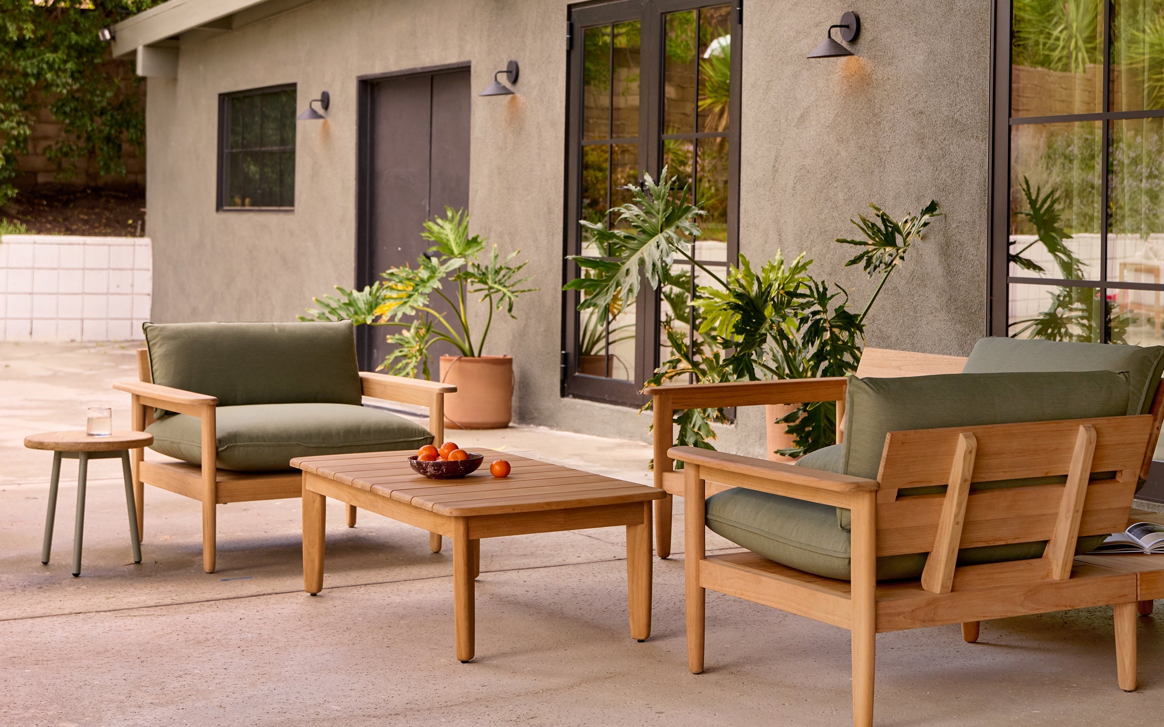 Outdoor patio furniture set with wooden frame, green cushions, a coffee table with fruit bowl, small side table, and potted plants.