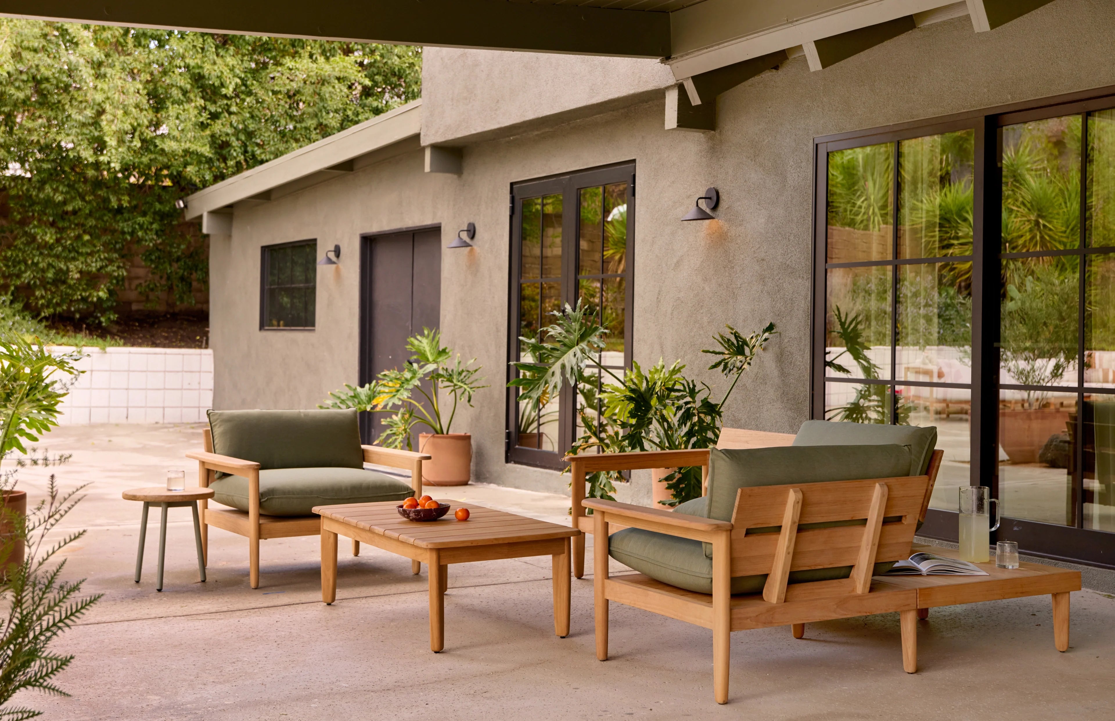 Outdoor patio seating set with wooden frame and green cushions, accompanied by a wooden coffee table, side table, and surrounding potted plants.