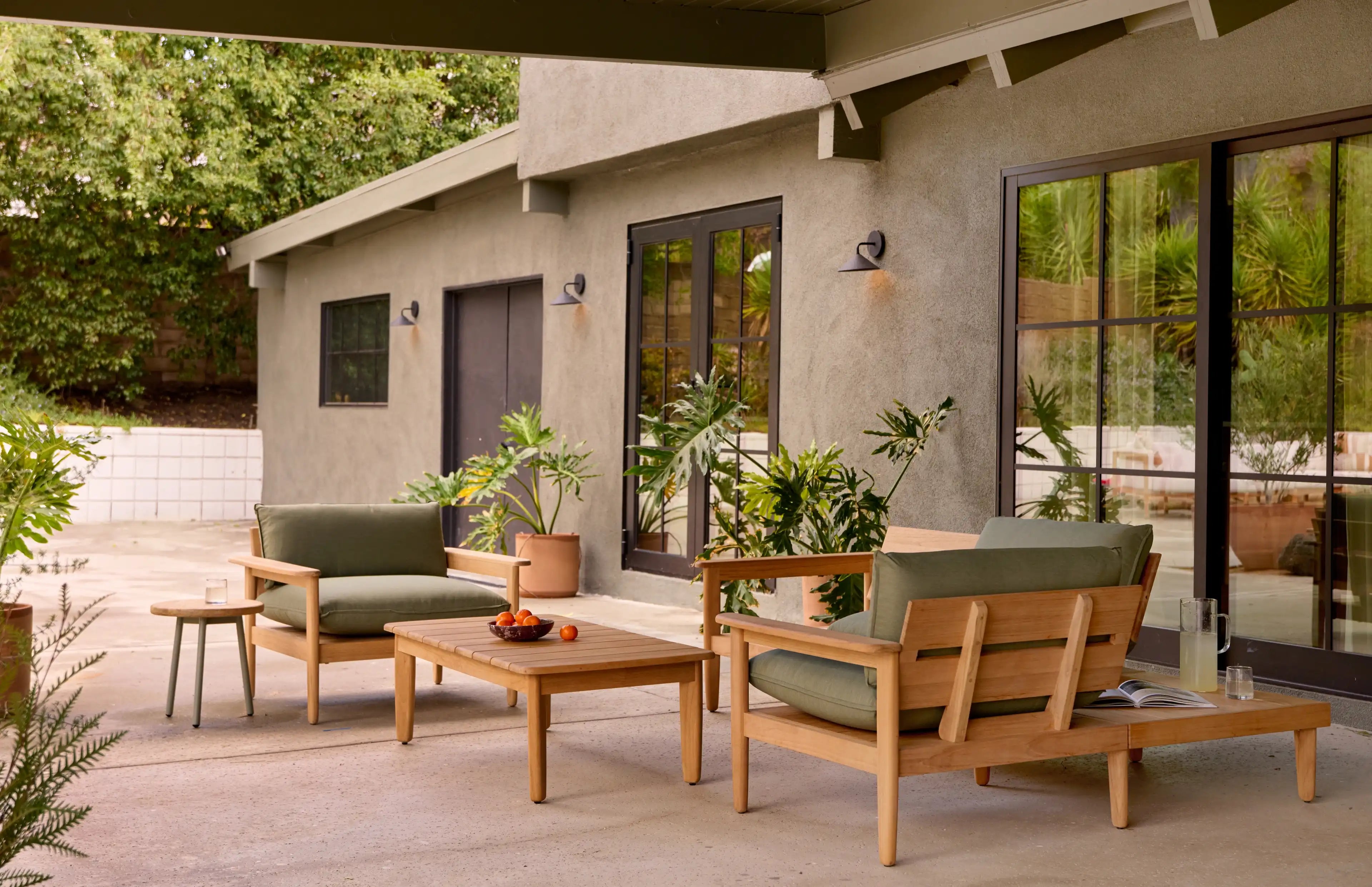 Outdoor patio set with wooden frame and green cushions, including two armchairs, a coffee table, and side table, surrounded by potted plants.