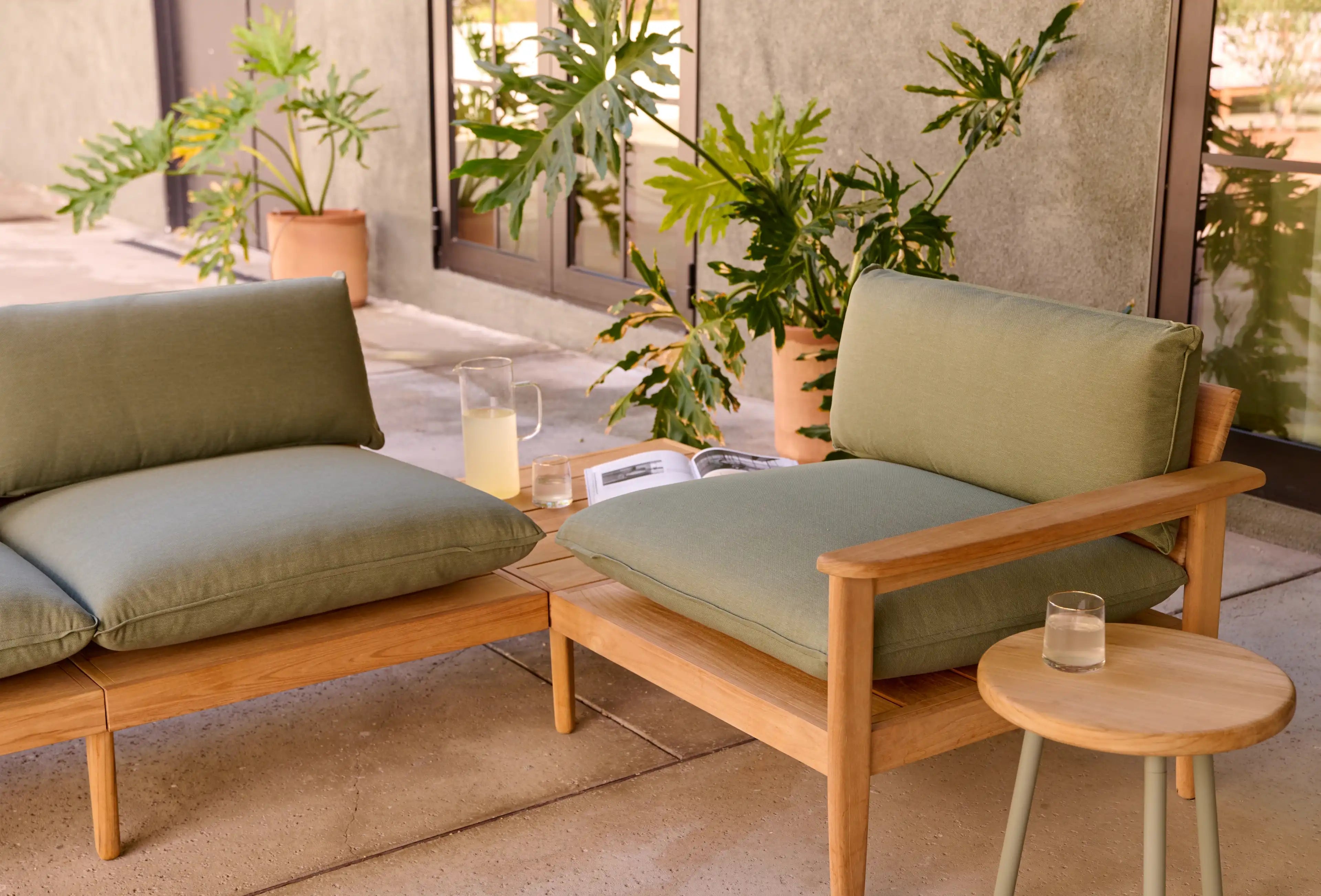 Outdoor wooden lounge chairs with green cushions set on a patio, accompanied by a small round wooden table and potted plants.