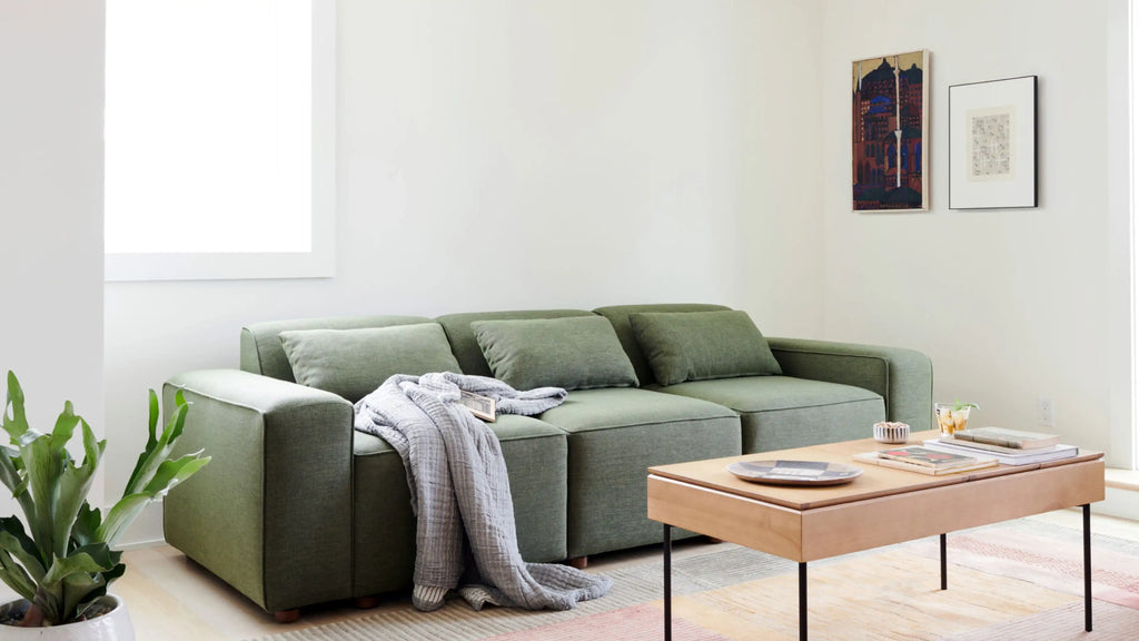 a green sectional sofa with pillows, blankets, and a wooden coffee table in a white living room with light hardwood flooring