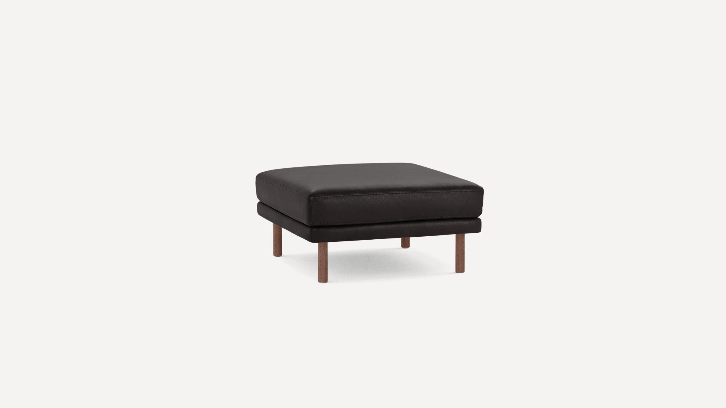 Square black leather ottoman with cushioned top and four wooden legs on a plain white background.