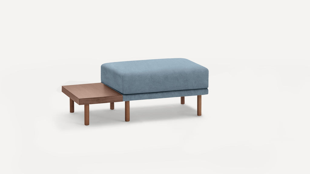 Modern ottoman with soft blue cushion and attached wooden side table, supported by cylindrical wooden legs, on a white background.