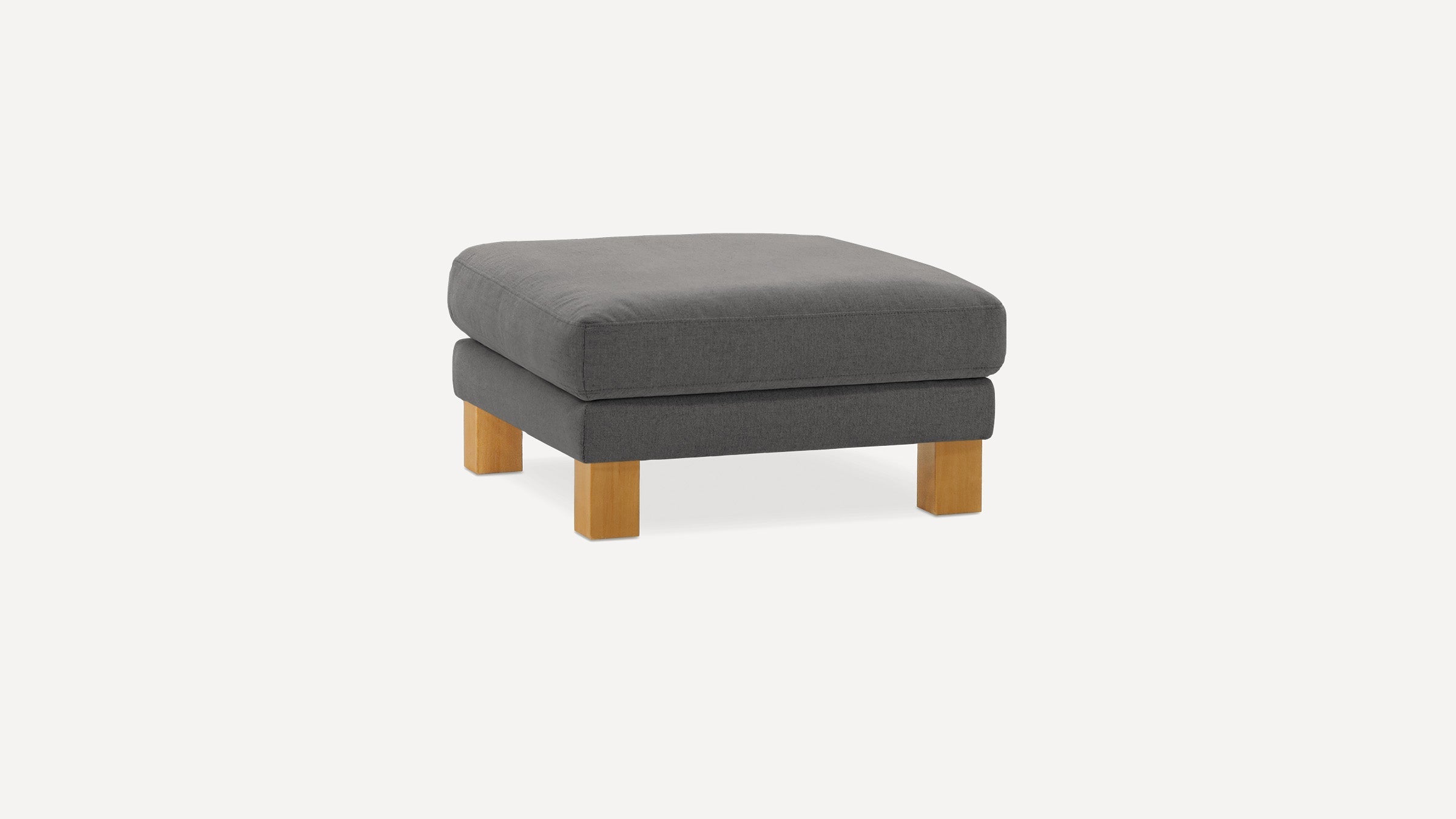 Square dark gray upholstered ottoman with thick cushion and four short wooden legs on white background.