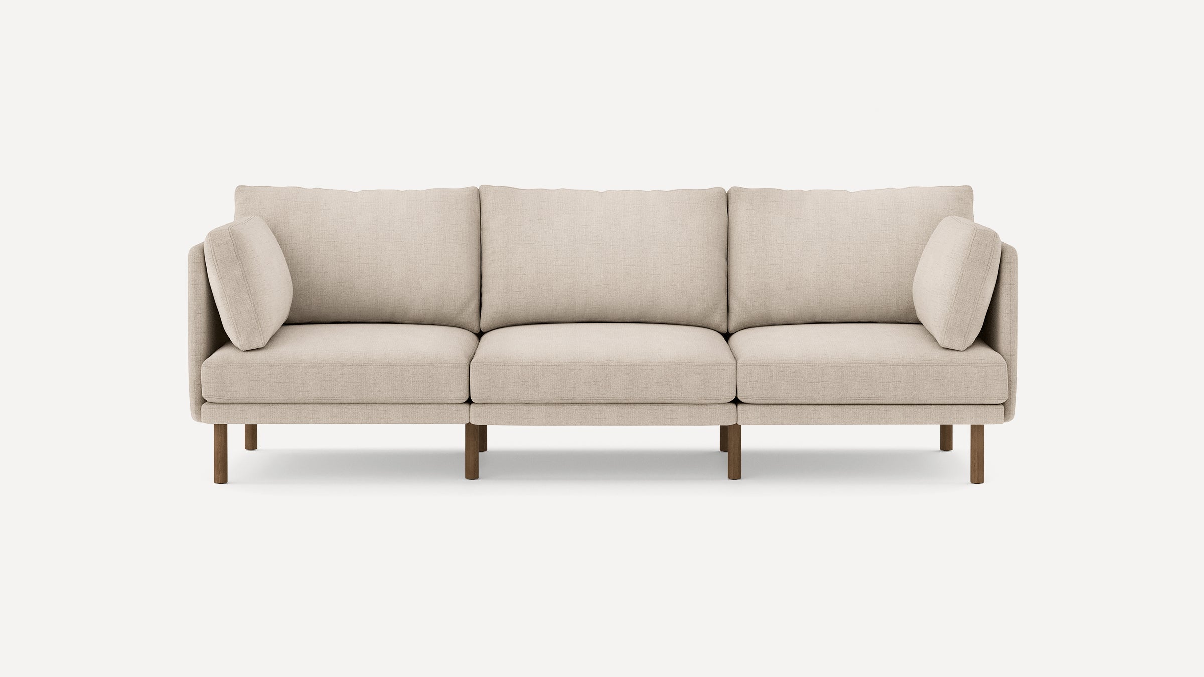 Modern beige three-seater sofa with fabric upholstery, three back cushions, two side pillows, and wooden legs on a white background.