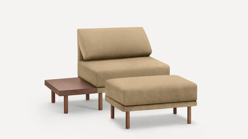 Beige upholstered armless lounge chair with matching ottoman and attached wooden side table, set on wooden legs.