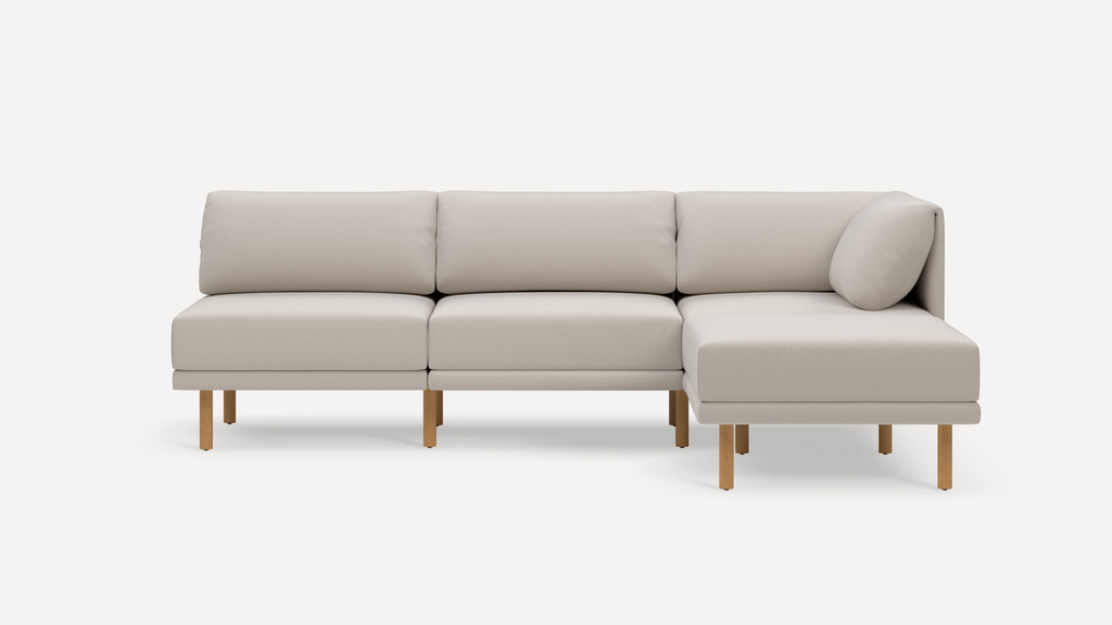 Beige L-shaped sectional sofa with wooden legs, three back cushions, and a side bolster pillow on the right end.