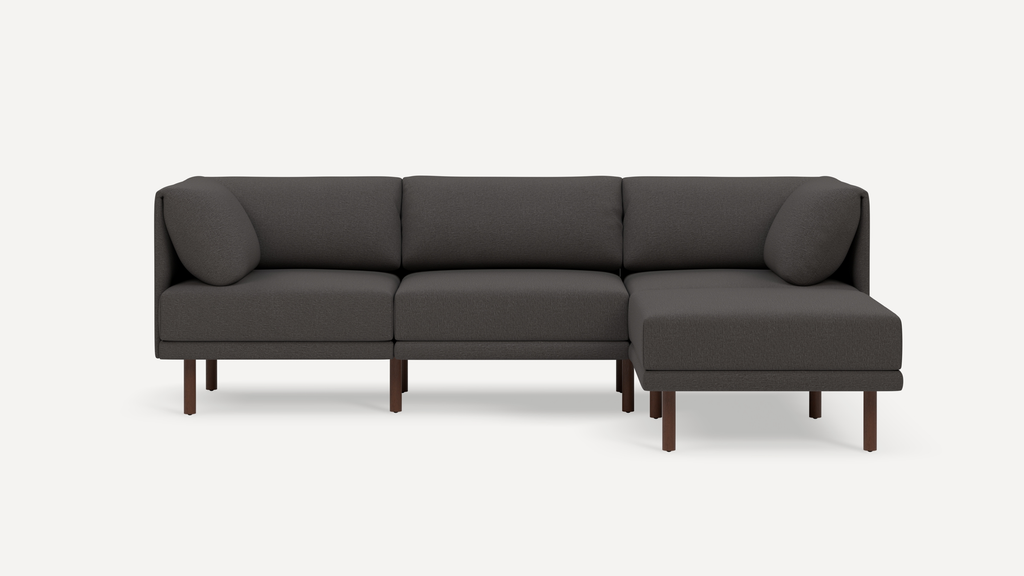 Modern dark gray fabric sectional sofa with wooden legs, featuring three seat cushions and two bolster pillows on a white background.