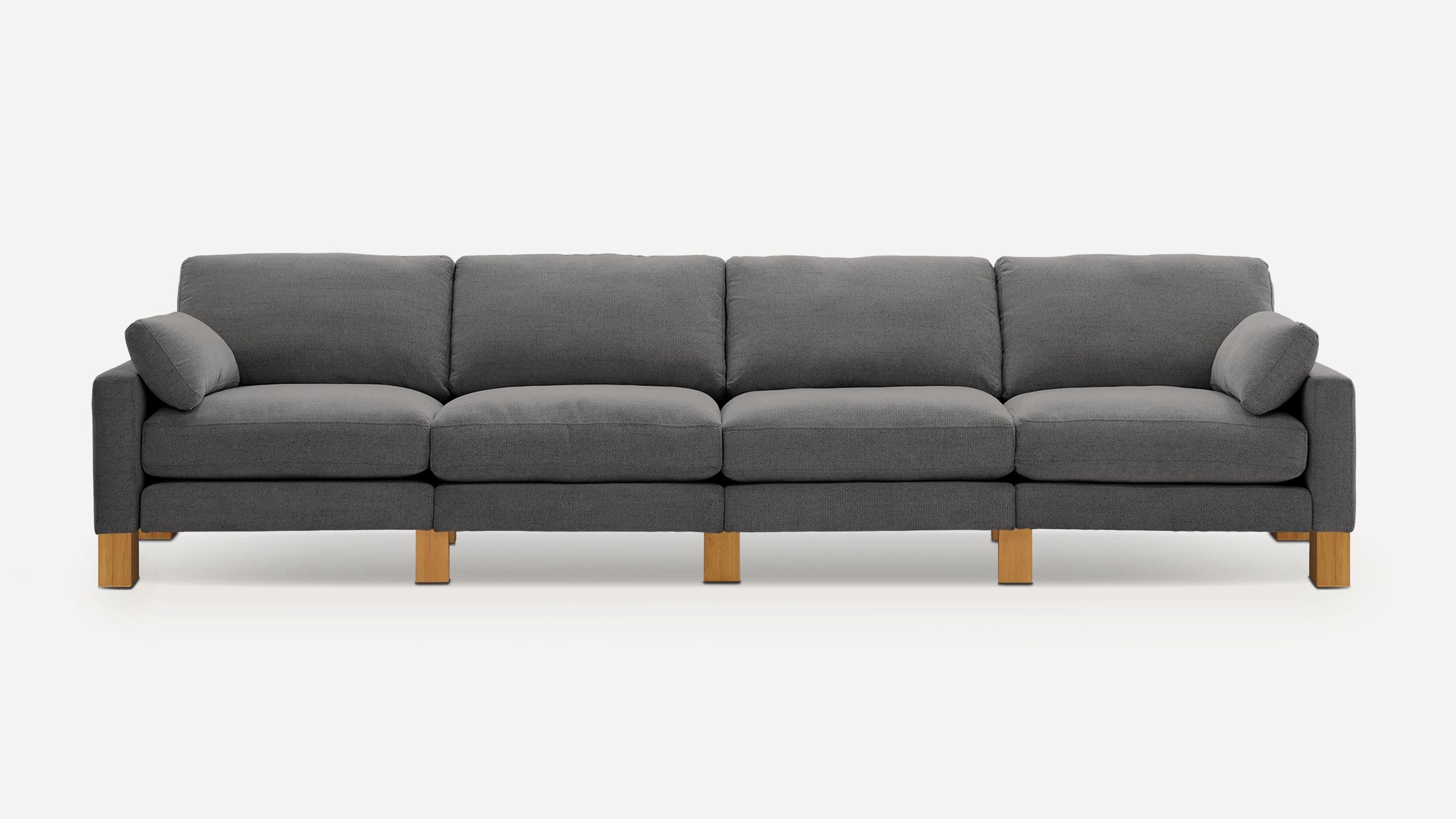 Long gray fabric sofa with four seat cushions, three back cushions, two accent pillows, and wooden block legs on plain background.