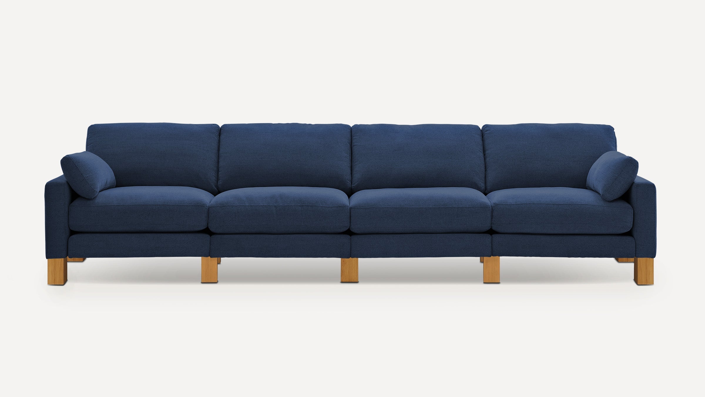 Modern four-seater sofa with deep blue fabric upholstery, plush cushions, two side pillows, and natural wooden block legs.