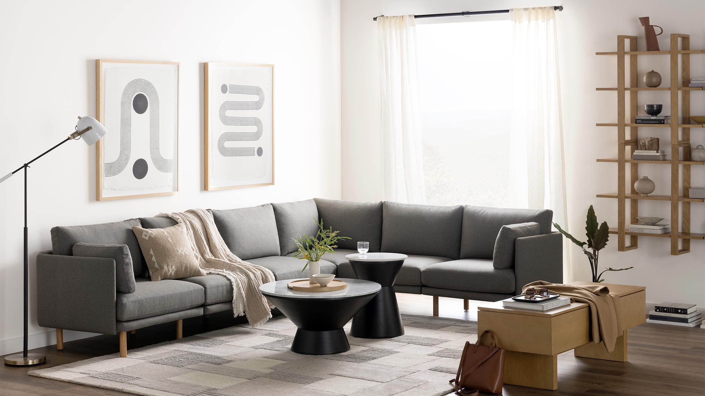 Modern living room with gray sectional sofa, black marble-top side tables, beige rug, wooden shelving unit, and large window with sheer curtains.