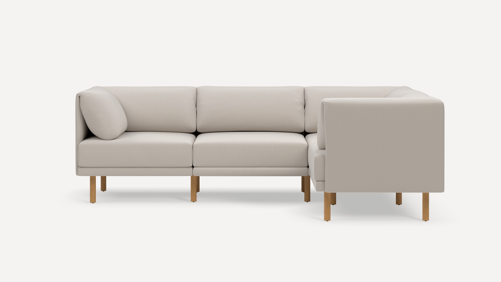 Light beige L-shaped sectional sofa with cushioned back, wooden legs, and a single round accent pillow on left side.