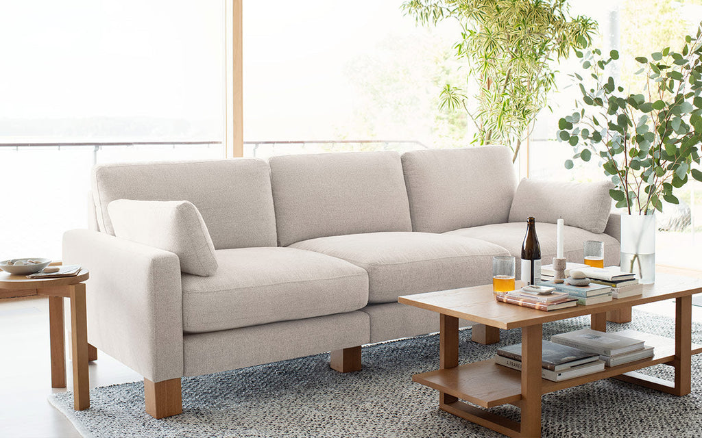 Light beige upholstered three-seater sofa with wooden legs in a bright living room, complemented by wooden coffee and side tables.