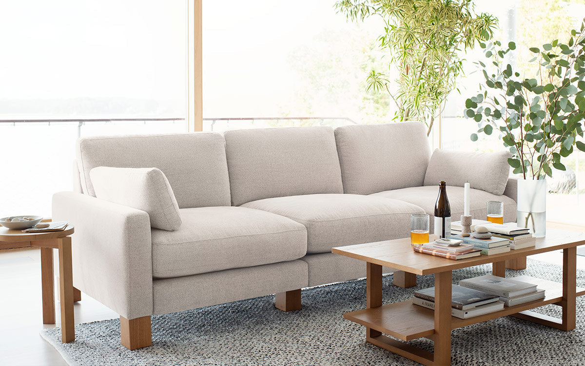 Light beige upholstered three-seater sofa with wooden legs in a bright living room, complemented by wooden coffee and side tables.