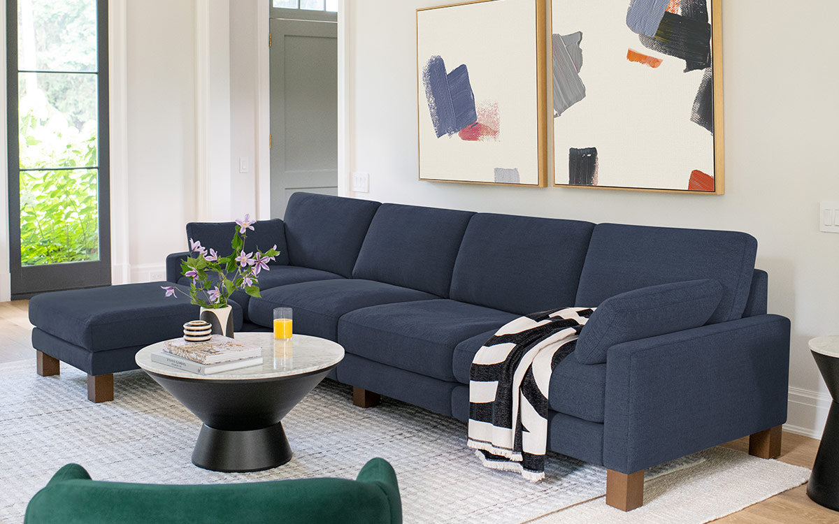 Modern navy blue sectional sofa with chaise lounge, wooden legs, black and white throw, and round marble coffee table in bright living room.