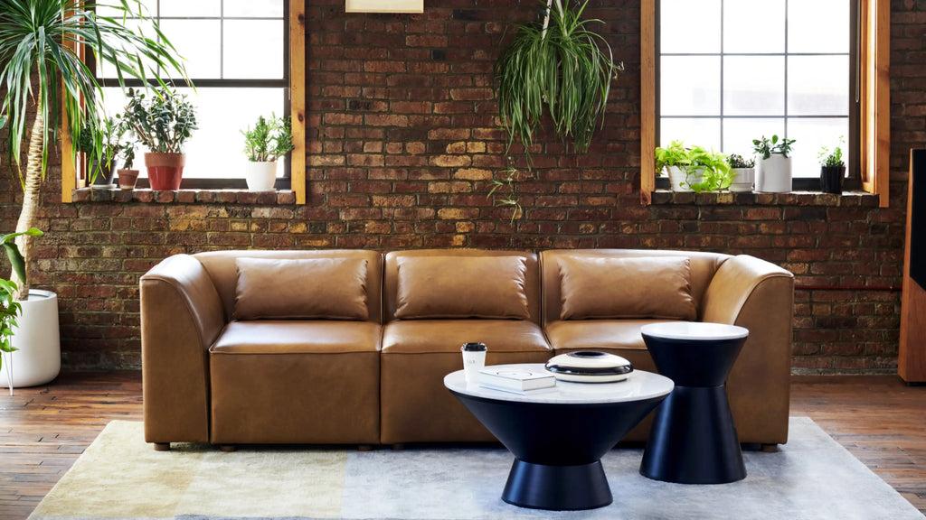 modern brown leather sectional sofa with black metal legs and matching round side tables in front of brick wall