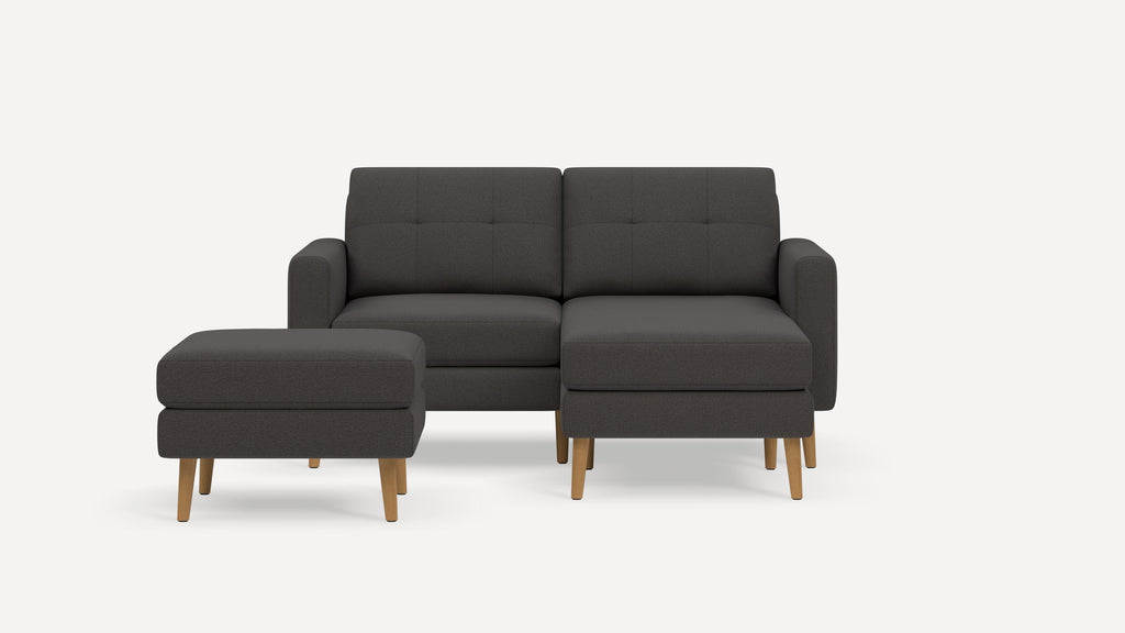 Dark gray upholstered two-seater sofa with wooden legs, tufted back cushions, and matching ottoman on white background.