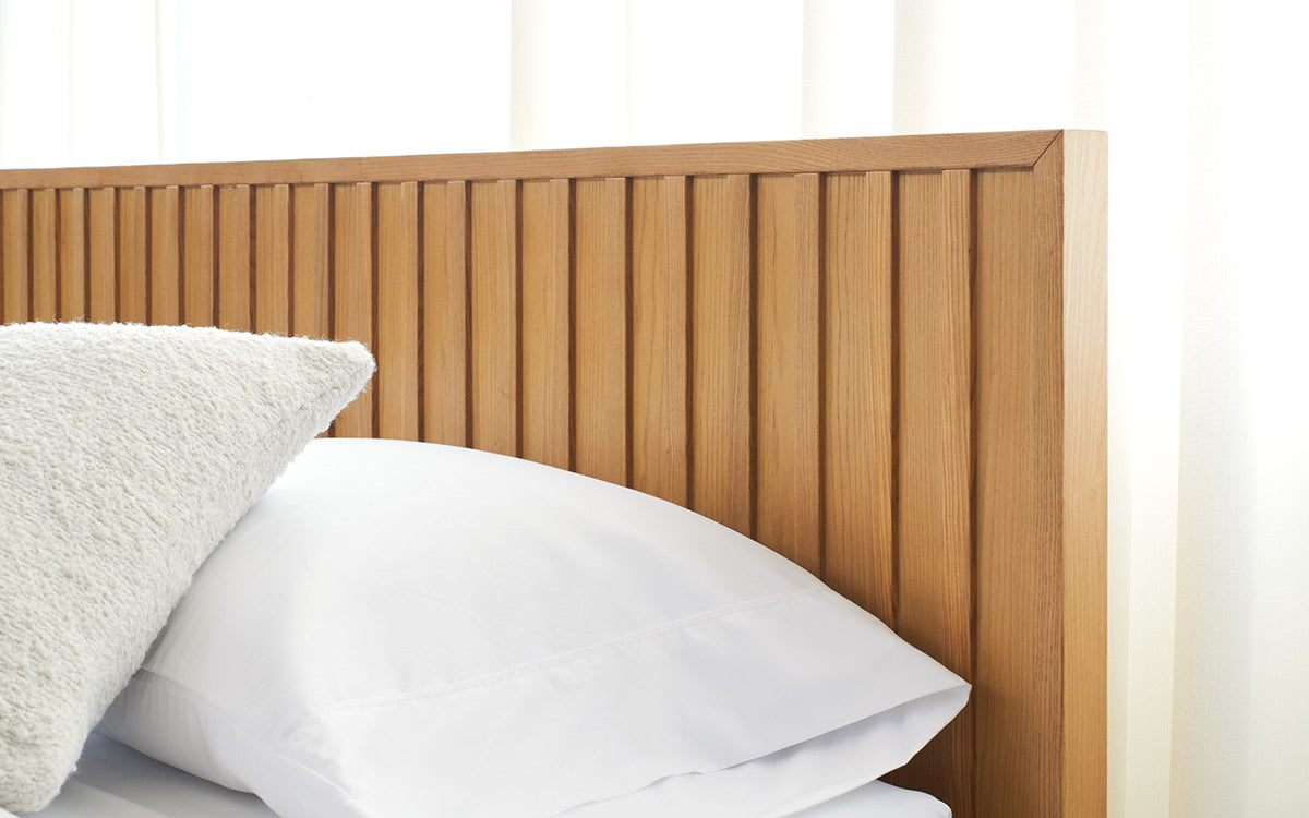 Close-up of a modern wooden slatted bed headboard with white pillows and a textured cream cushion against sheer white curtains.
