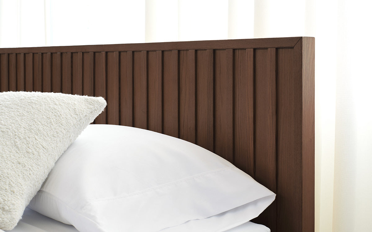 Close-up of a dark brown wooden slatted headboard with white bed pillows against sheer white curtains.