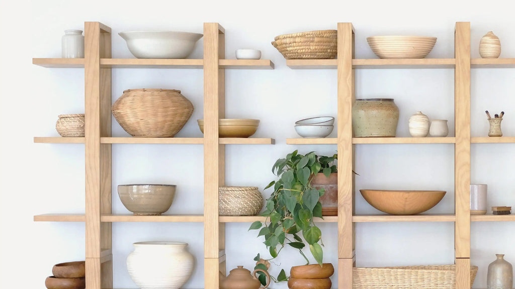 Light wooden shelving unit with assorted ceramic bowls, woven baskets, and green potted plant on a white wall background.