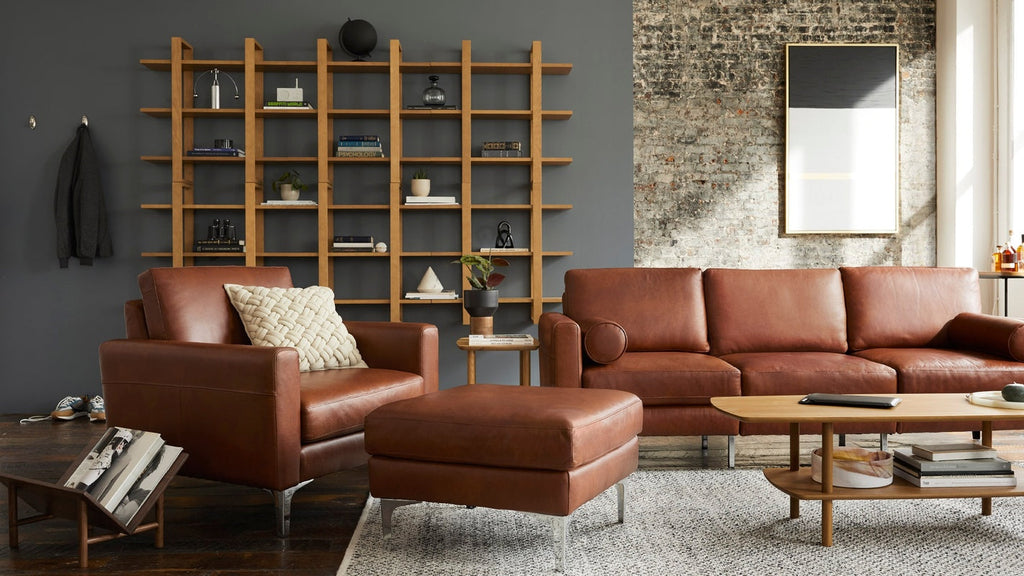Modern living room with cognac leather armchair, matching ottoman and sofa, wooden coffee table, and minimalist shelving.