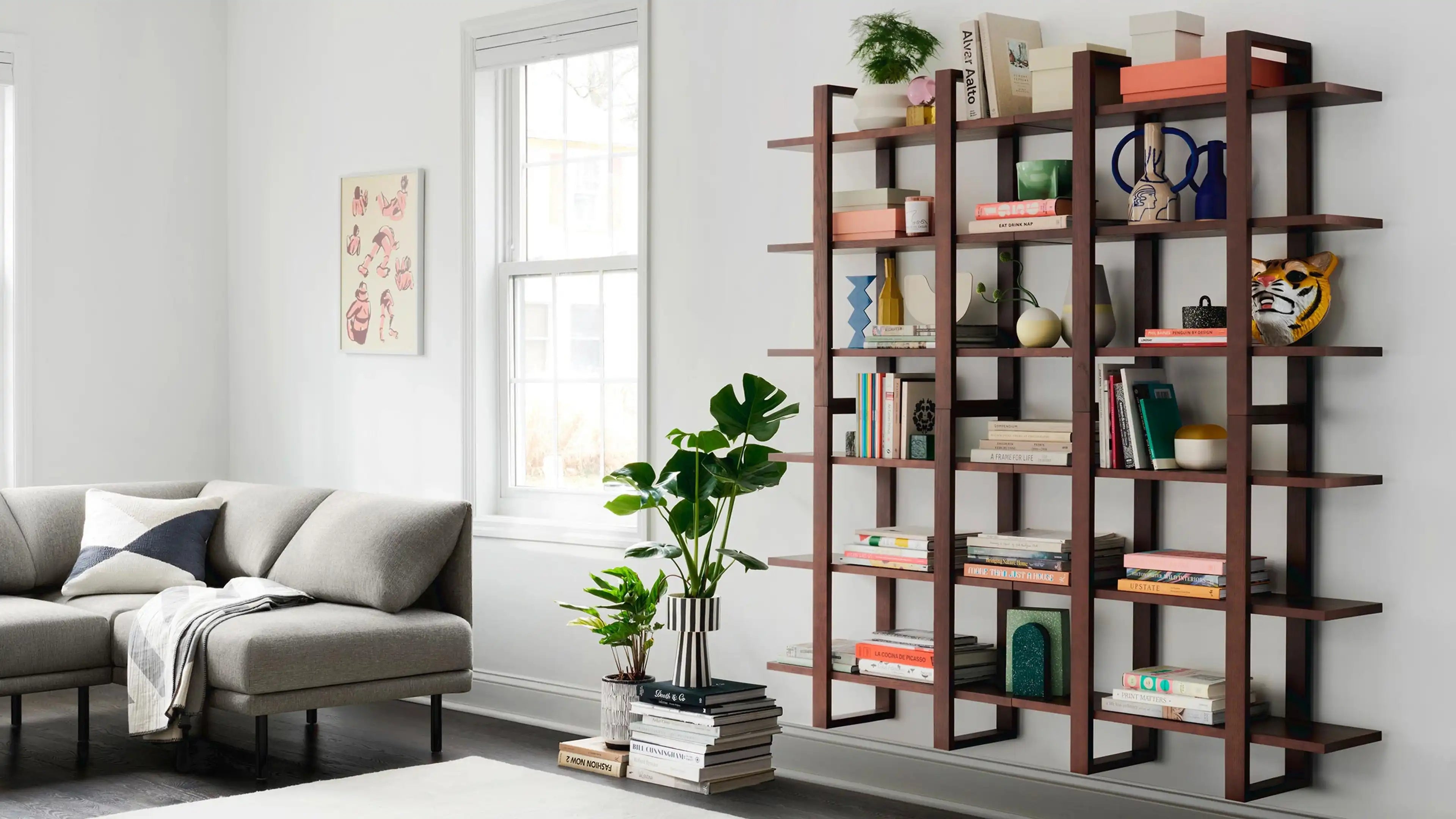Modern living room with a gray sectional sofa, open wood bookshelf filled with books and decor, and potted plants near a window.