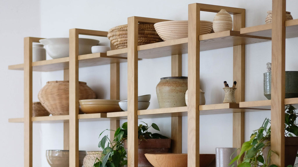 Light wooden open shelving unit displaying various ceramic bowls, vases, woven baskets, and green potted plants against a white wall.