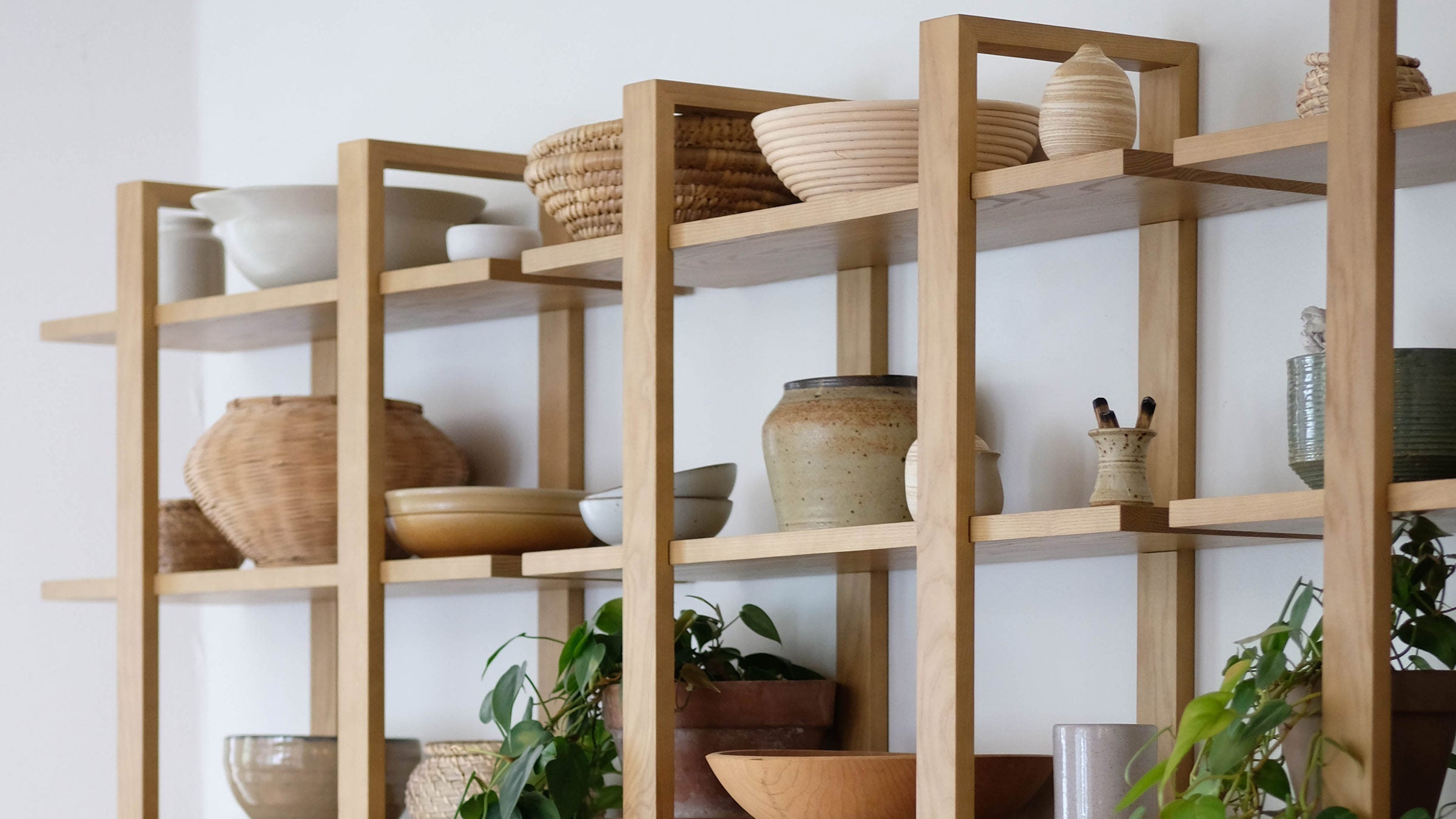 Wooden open shelving unit displaying ceramic pots, woven baskets, wooden bowls, and potted green plants in a minimalist setting.