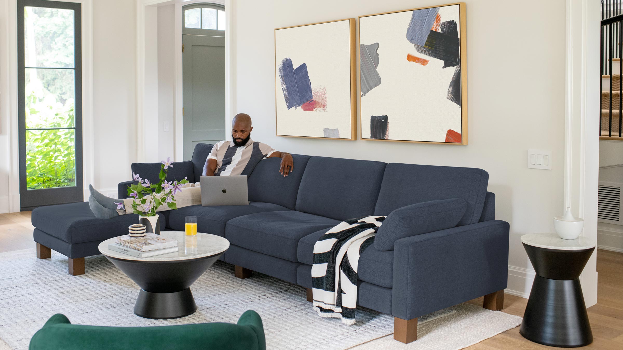 Man sitting on a dark blue sectional sofa with wooden legs in a modern living room featuring abstract wall art and round marble-top tables.