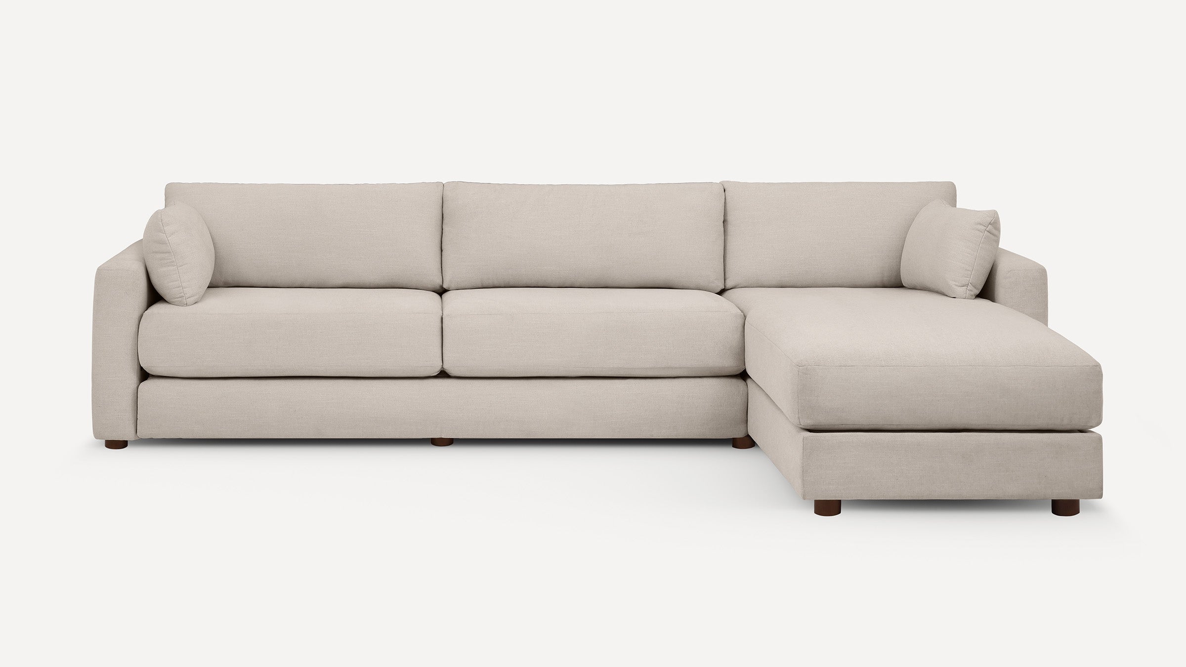 Beige sectional sofa with chaise lounge, three back cushions, two armrest pillows, and wooden legs on a white background.