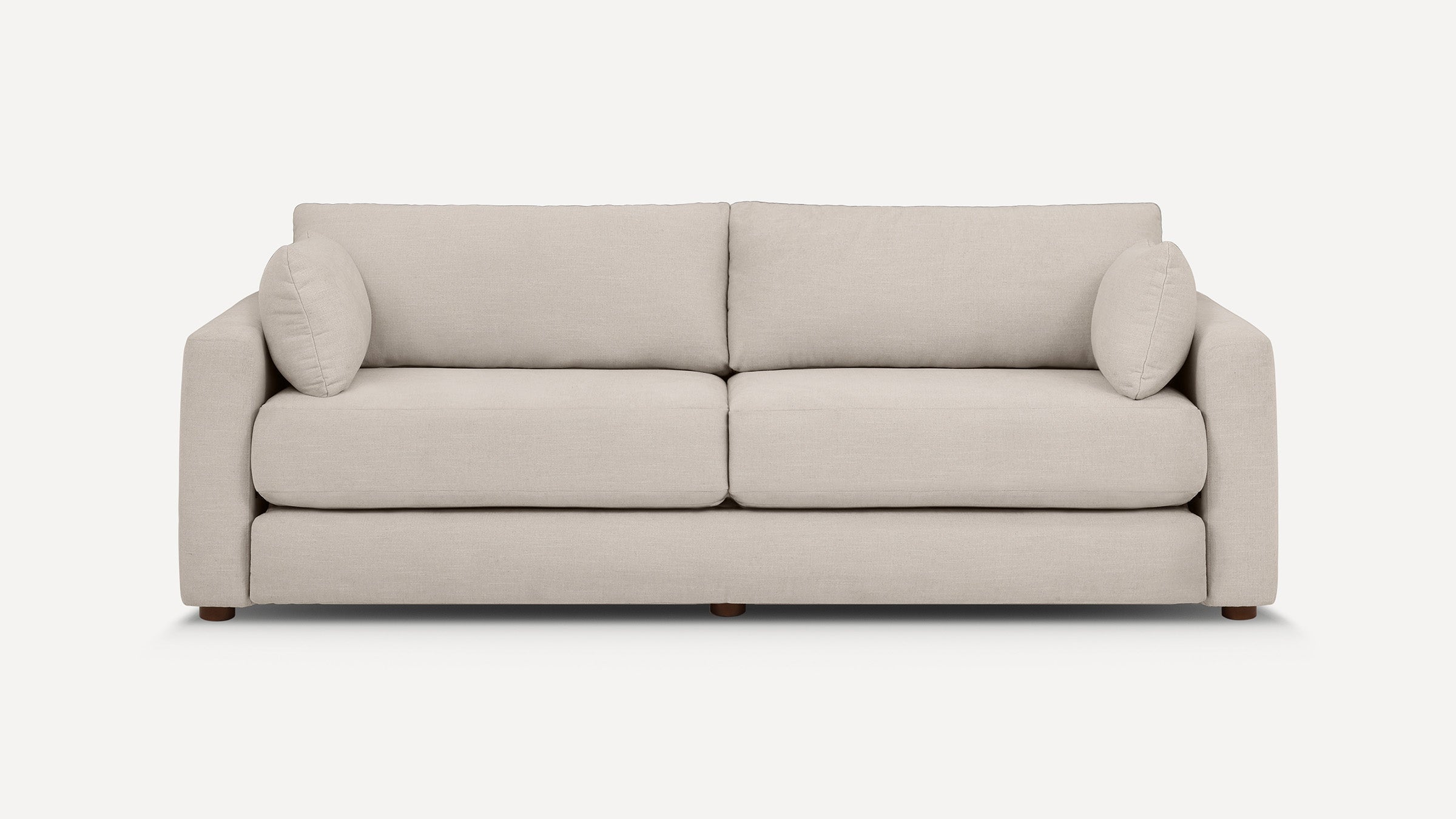 Modern beige fabric three-seater sofa with cushioned backrest, armrests, and two round throw pillows, set on small wooden legs.