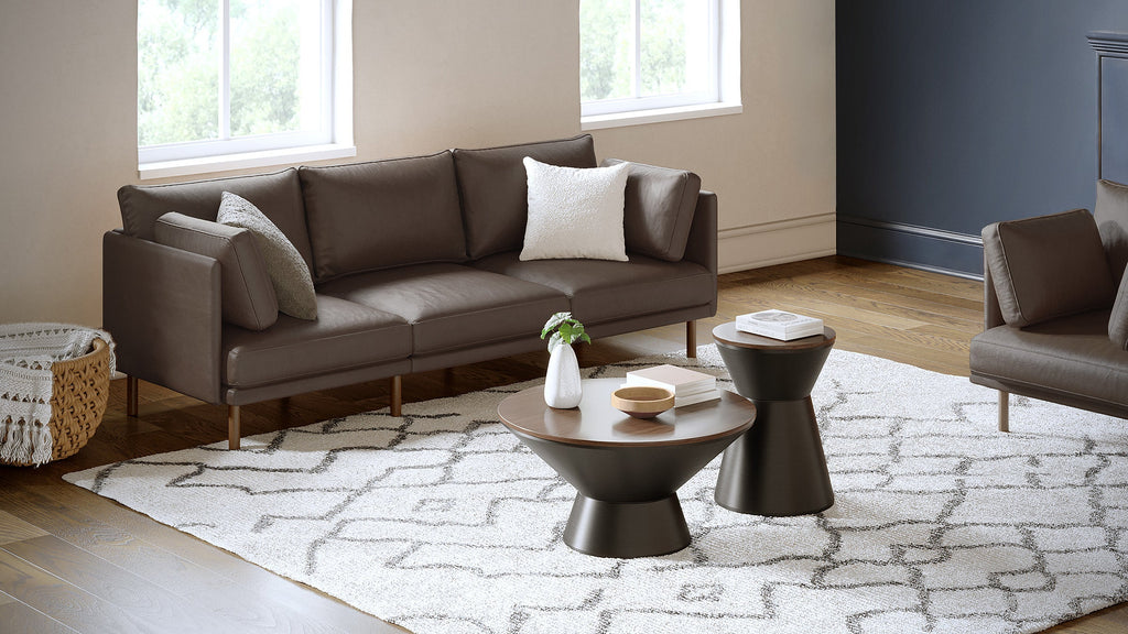Modern living room featuring a brown leather sofa with pillows, two matching hourglass-shaped coffee tables, a patterned rug, wooden floor, and natural light from windows.