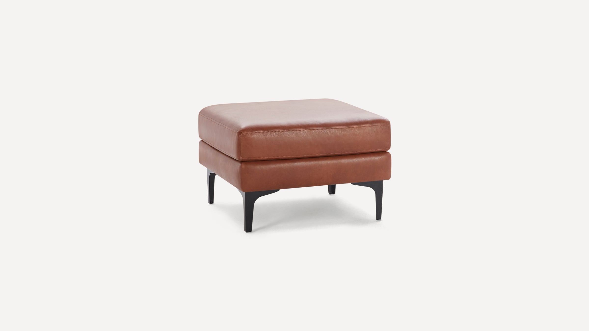 Square brown leather ottoman with thick cushioned top and black tapered metal legs on a white background.
