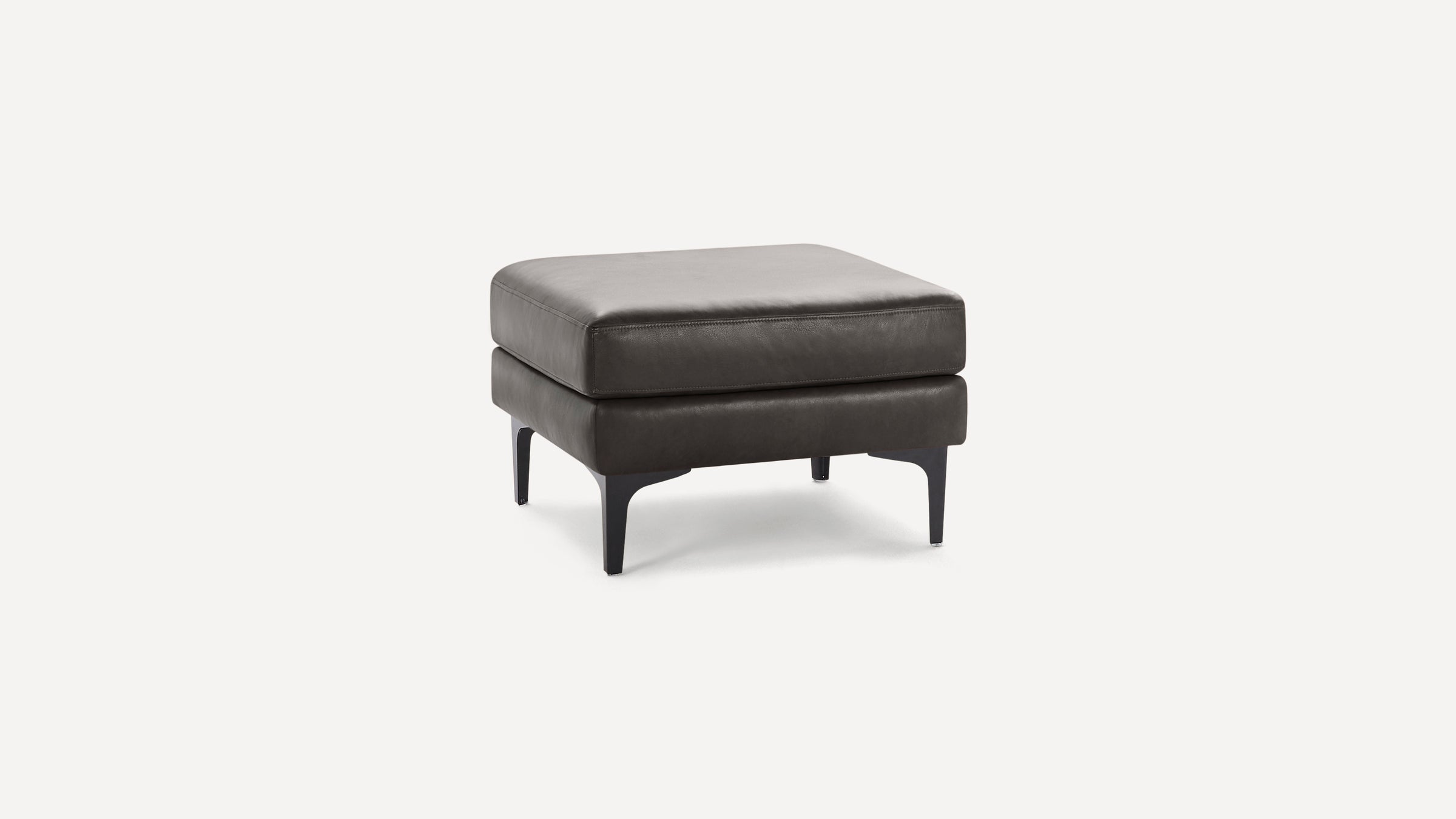 Square dark gray leather ottoman with cushioned top and four slender black legs on a white background.