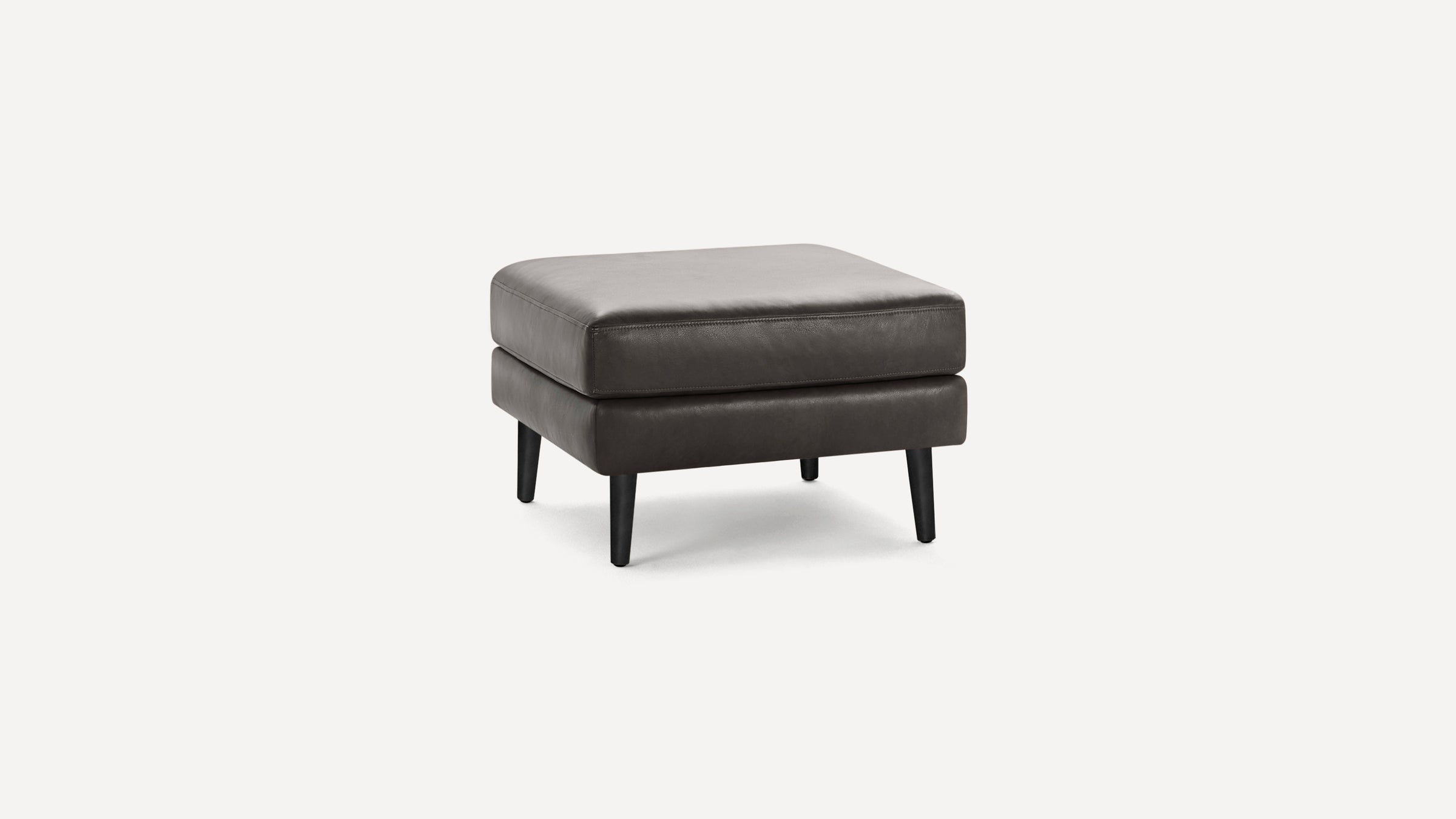 Square dark gray leather ottoman with black tapered wooden legs on a white background.