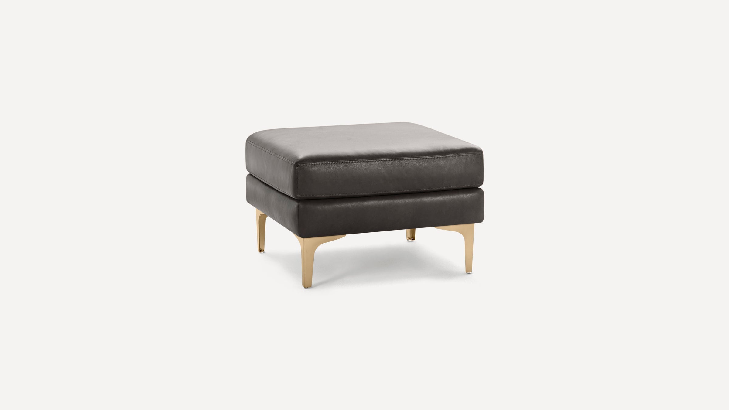 Square dark gray leather ottoman with thick cushioning and gold metal legs on a white background.